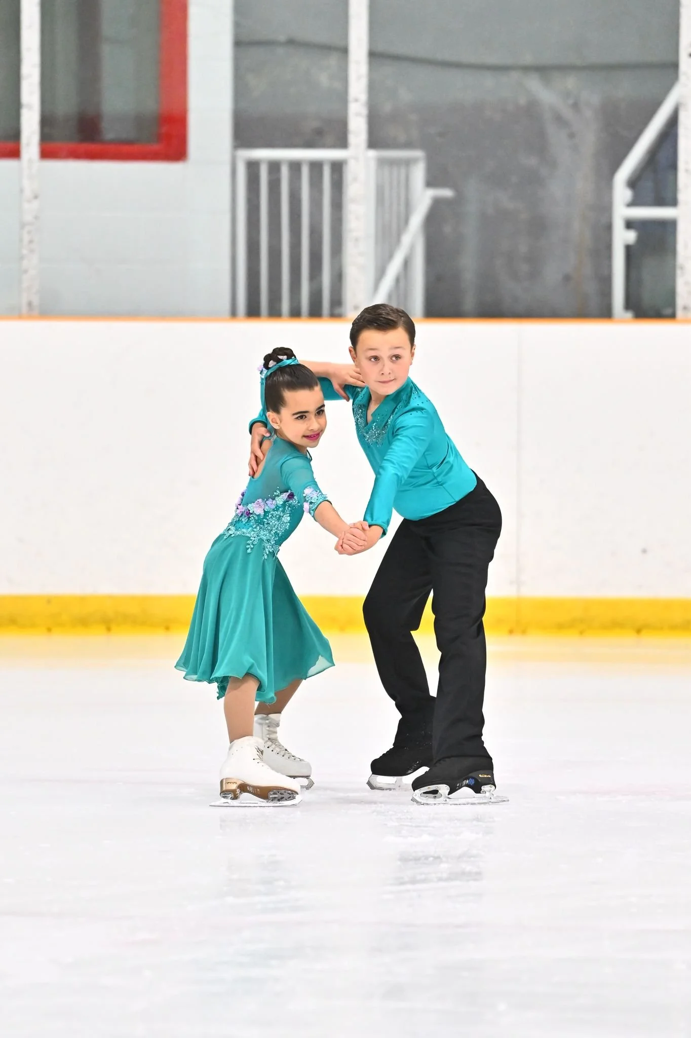 A young girl and boy figure skating on ice rink, holding hands and looking to the side, dressed in bright blue outfits.