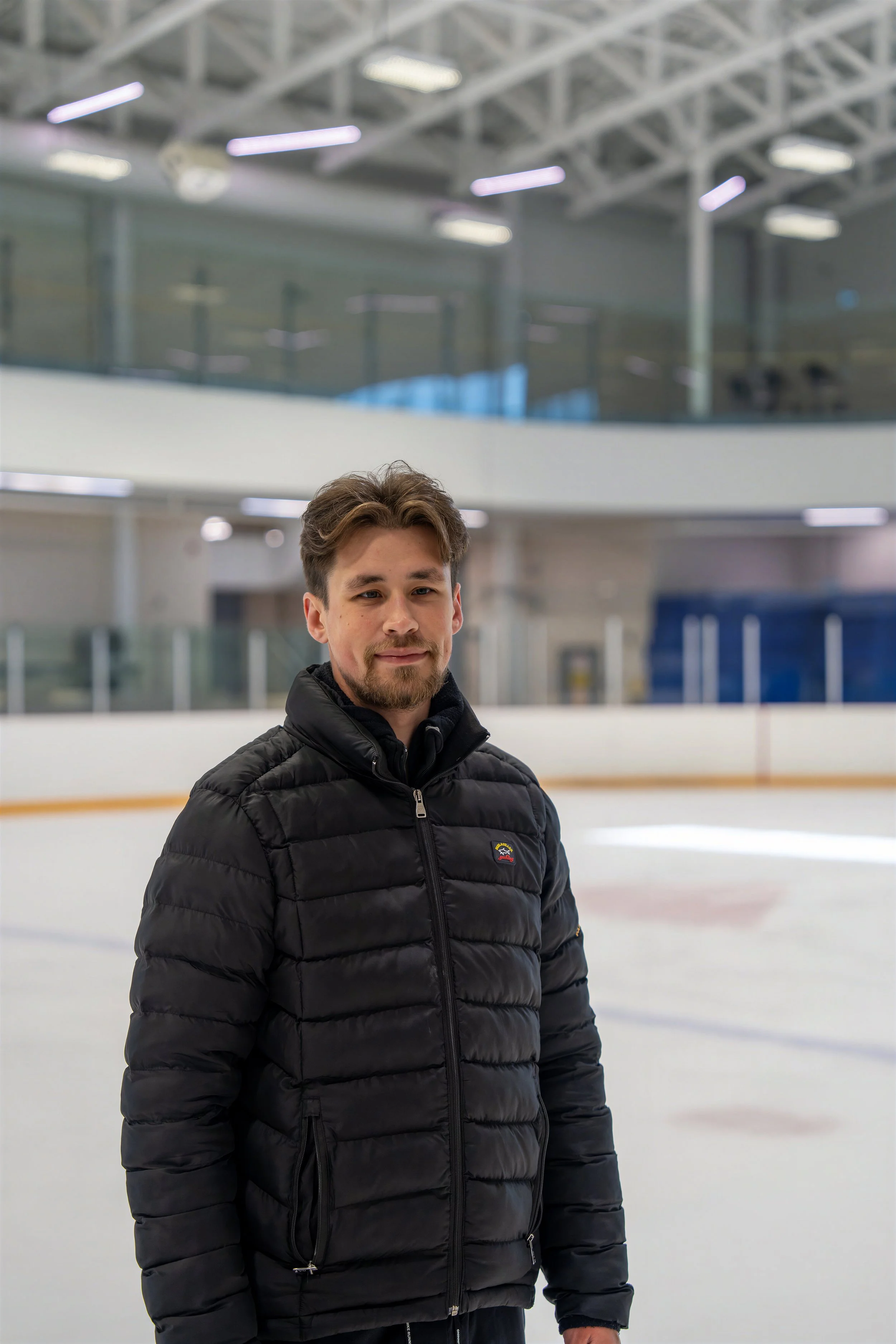 A young man with brown hair and a beard wearing a black puffer jacket and standing on an indoor ice rink.