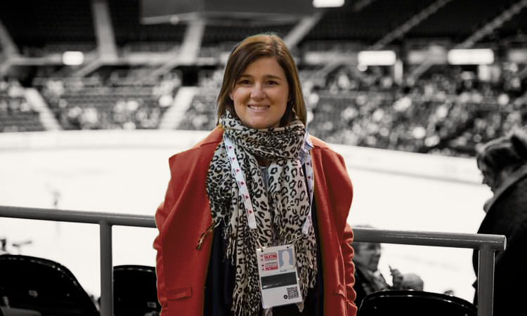 A smiling woman at an ice skating rink, wearing a red coat and leopard print scarf, with a credential badge hanging around her neck.