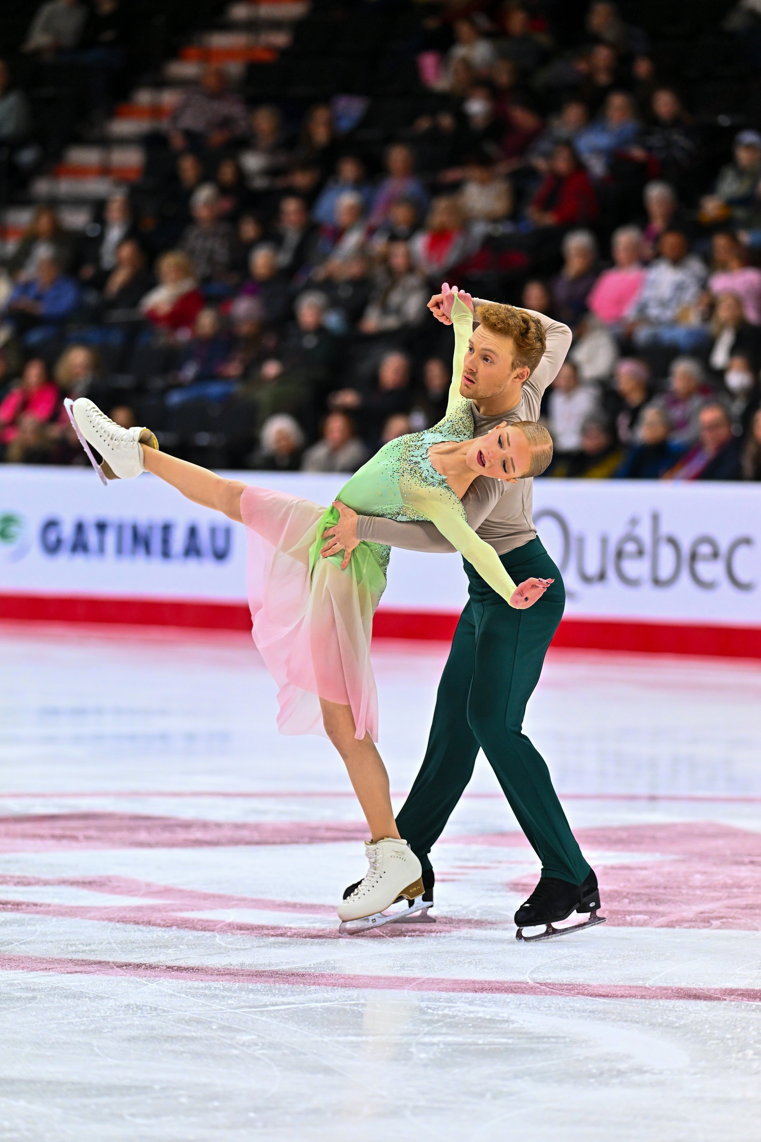 A figure skater performing a lift on the ice rink during a competition, with an audience in the background.