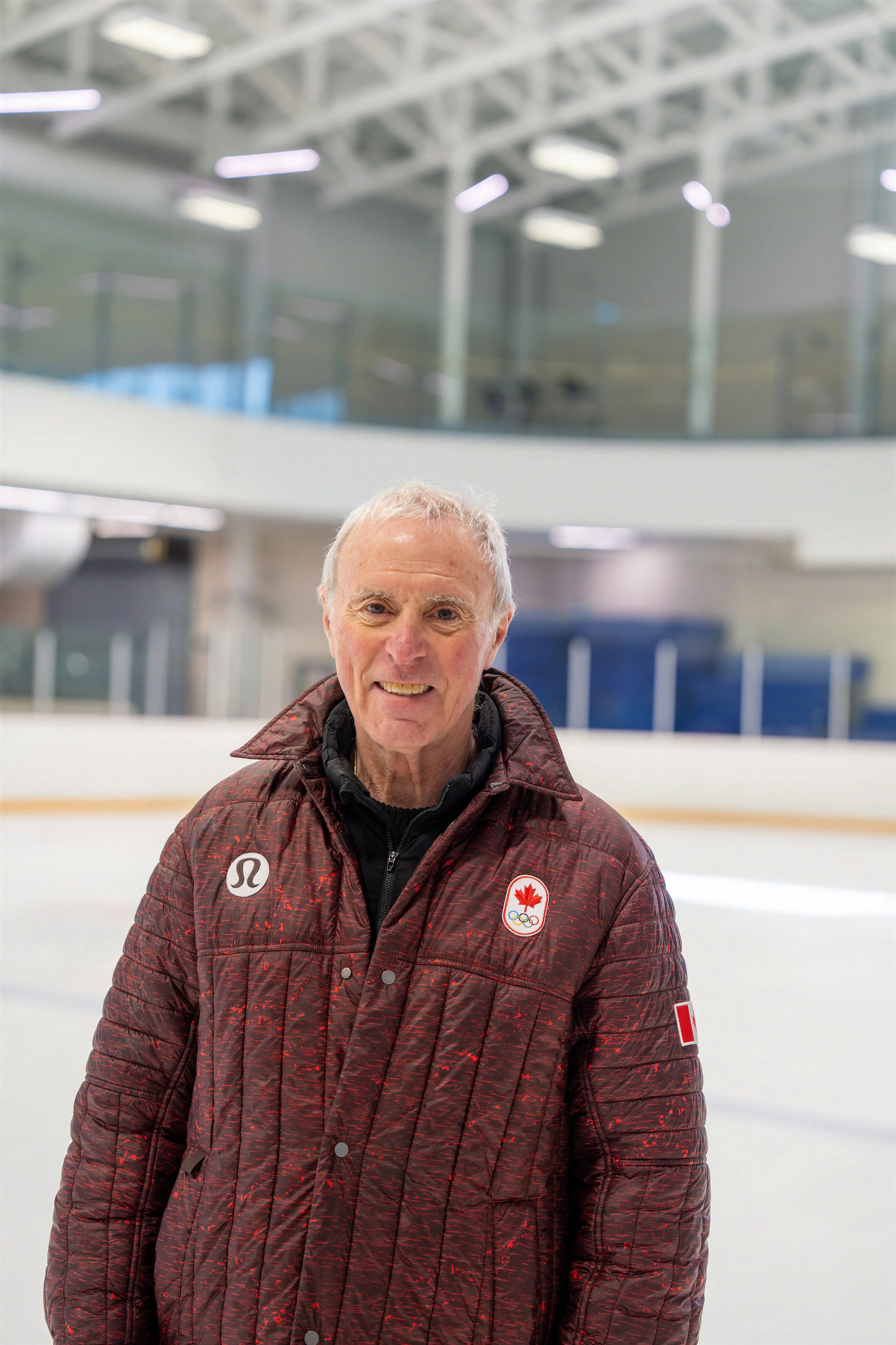 An older man standing on an ice hockey rink, wearing a maroon patterned Canada Olympic jacket with Canadian and Olympic logos, smiling at the camera.