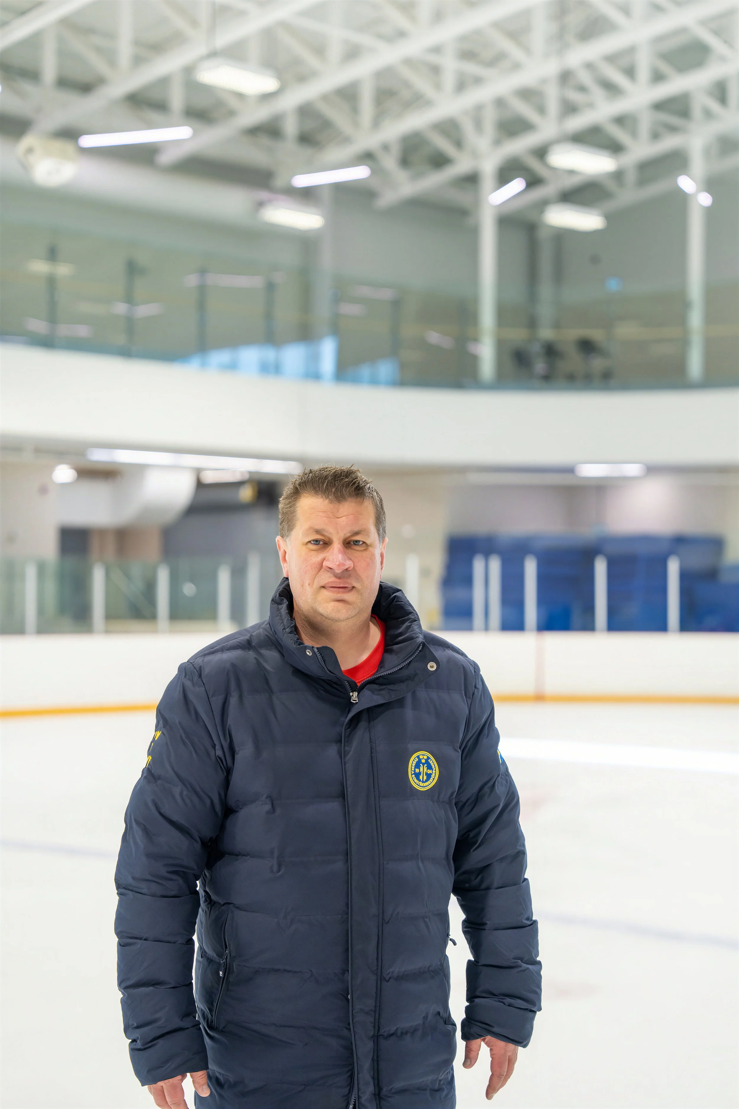 Man wearing a navy blue jacket with a yellow and blue emblem on the left chest, standing on an ice rink.