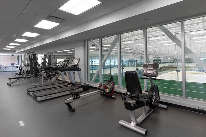 Empty gym with treadmills and exercise bikes near large glass windows overlooking an indoor sports rink.