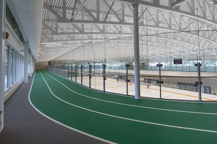 Interior of an indoor running track and ice rink, with green track surface, glass walls, and a modern, arched ceiling structure.