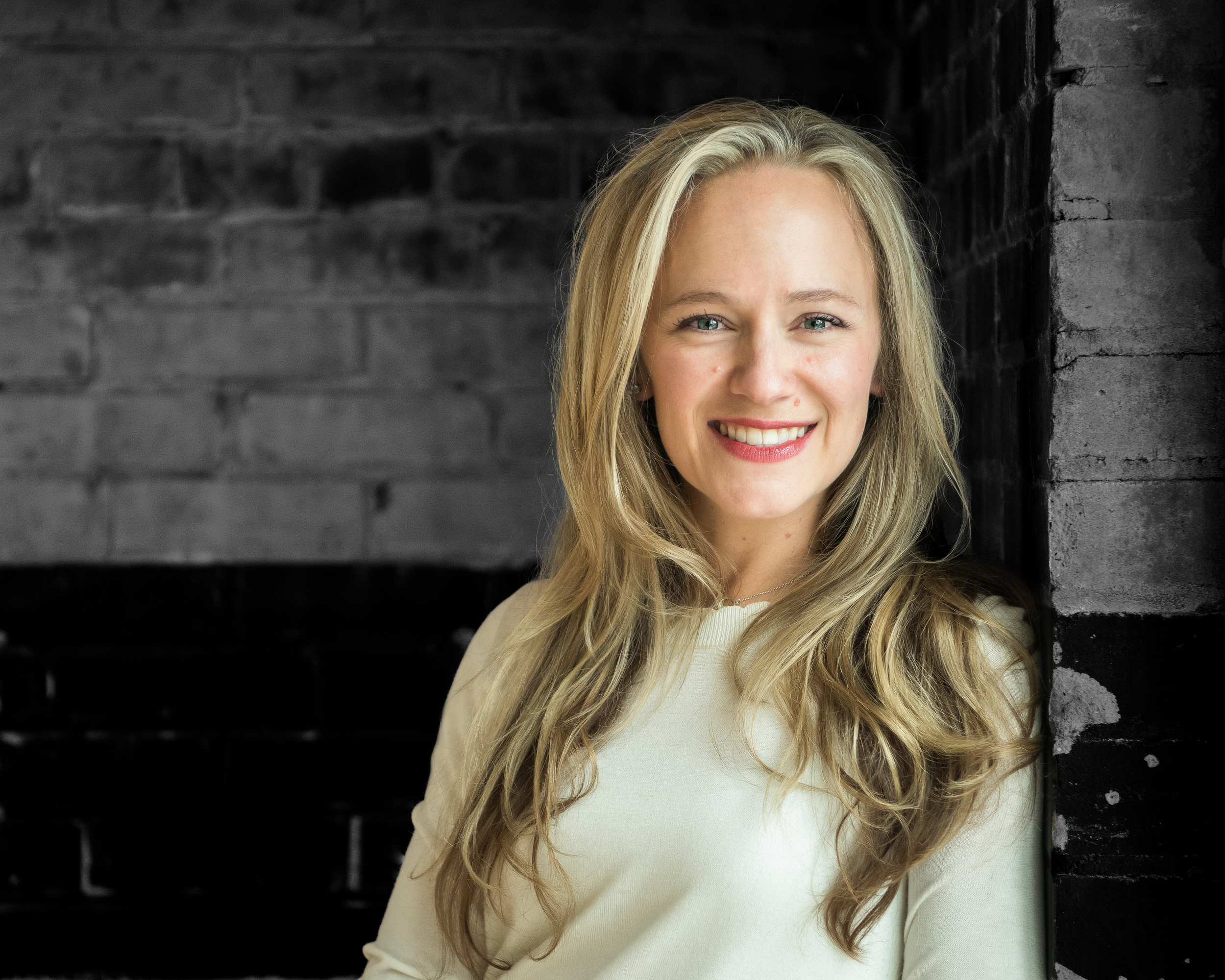 A smiling woman with long blonde hair wearing a white top, standing against a dark brick wall.