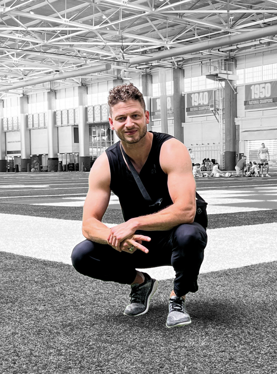A man with curly hair and a beard in athletic clothing squatting inside a large indoor sports facility with banners hanging from the high ceiling, people in the background, and a track surface underfoot.