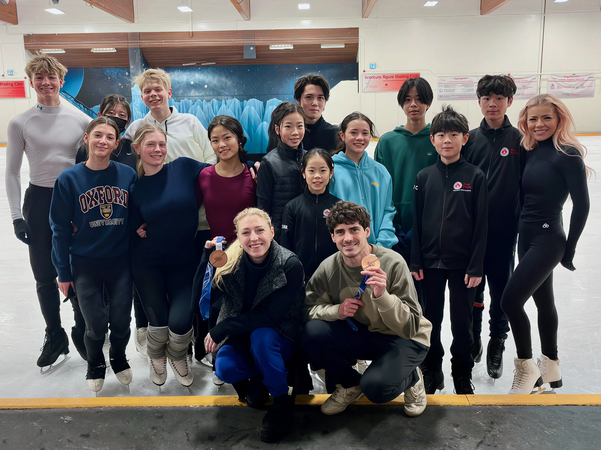 A group of young figure skaters, both boys and girls, posing together on ice rink with medals, some wearing warm-up jackets and skating gear, smiling for the photo. The background has an icy mural and a wall with signs.