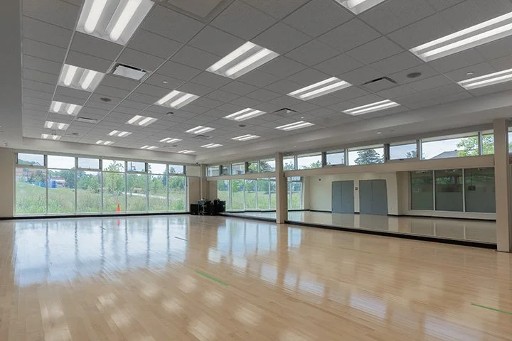 Empty gym or dance studio with wooden flooring and large windows, illuminated by ceiling lights.