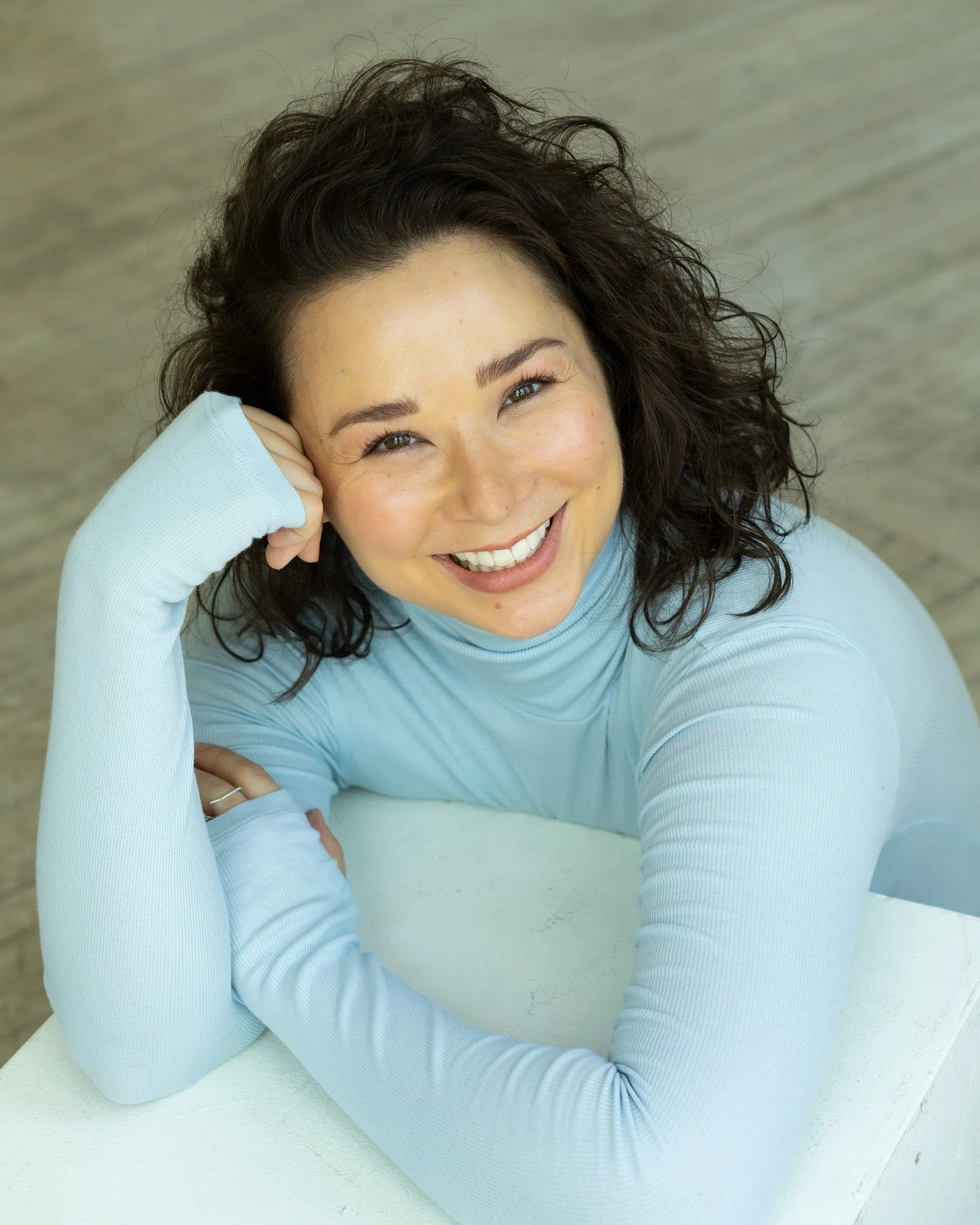A woman with curly brown hair resting her head on her hand, smiling while looking up, wearing a light blue long sleeve shirt, seated at a white table.