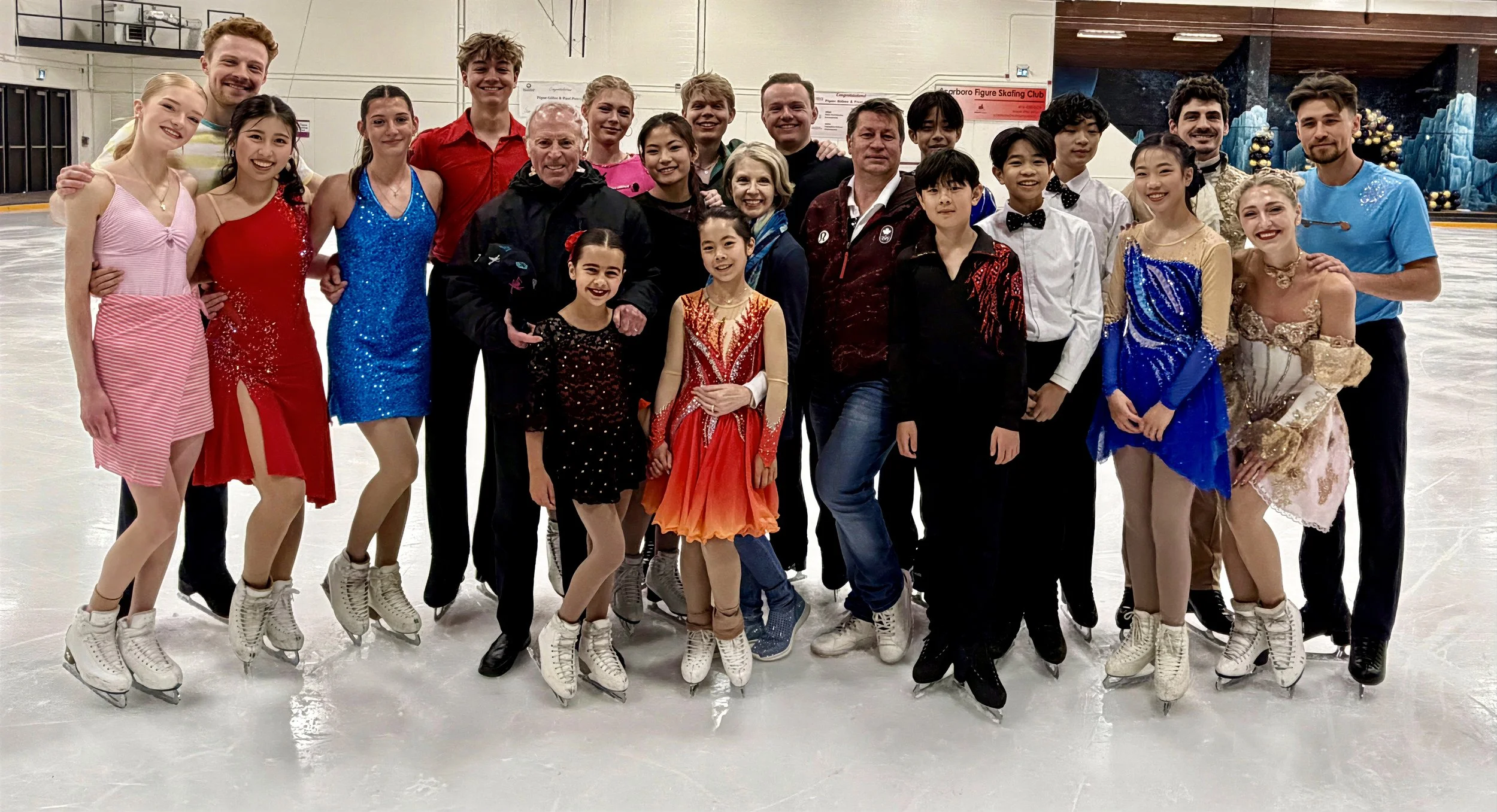 Group of ice skaters, including children, teenagers, and adults, posing on an ice rink after a skating event or practice, with some wearing colorful costumes and figure skates.