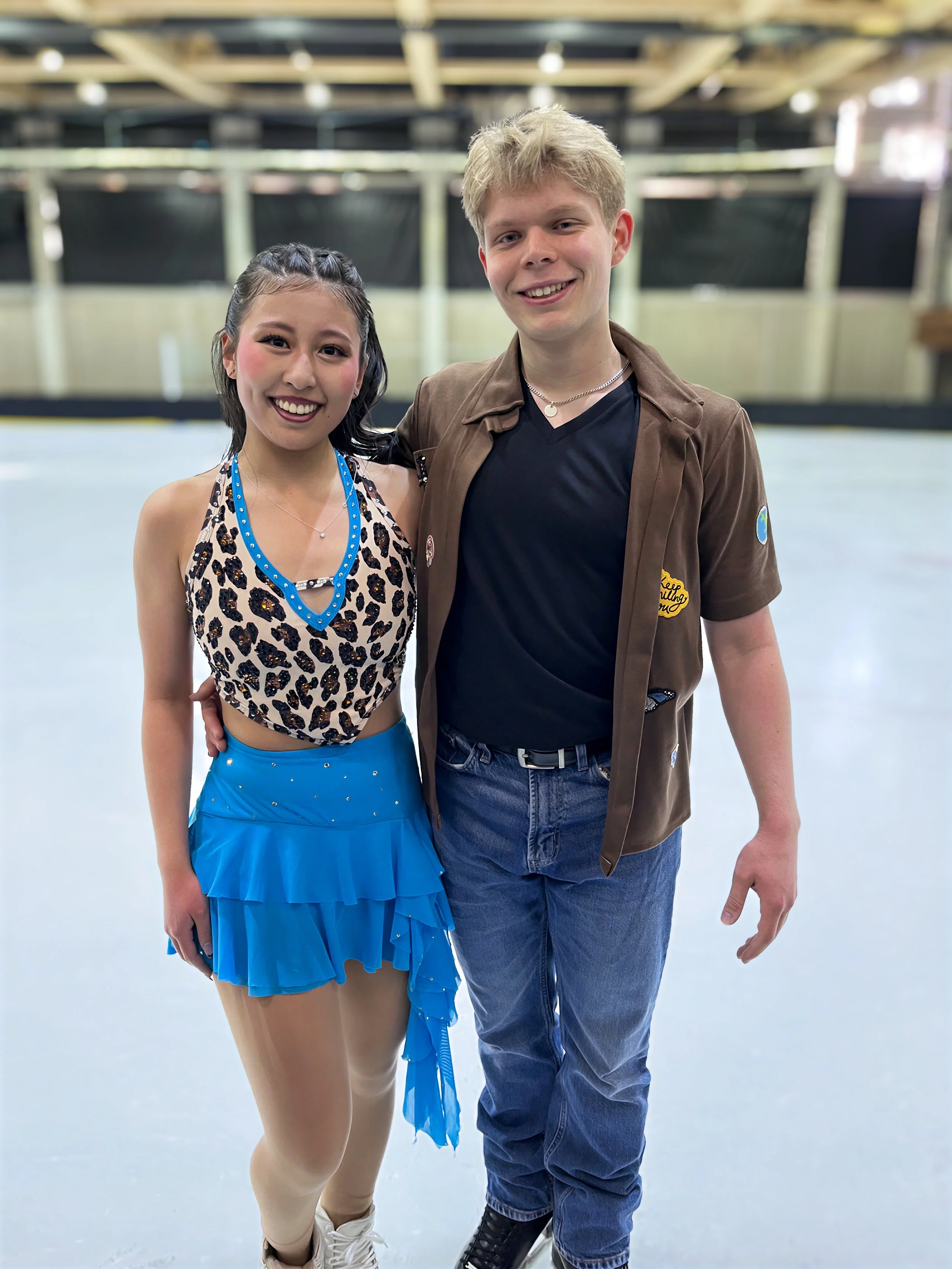A young woman in a blue figure skating dress and a young man in casual clothing pose on an ice rink, smiling at the camera.
