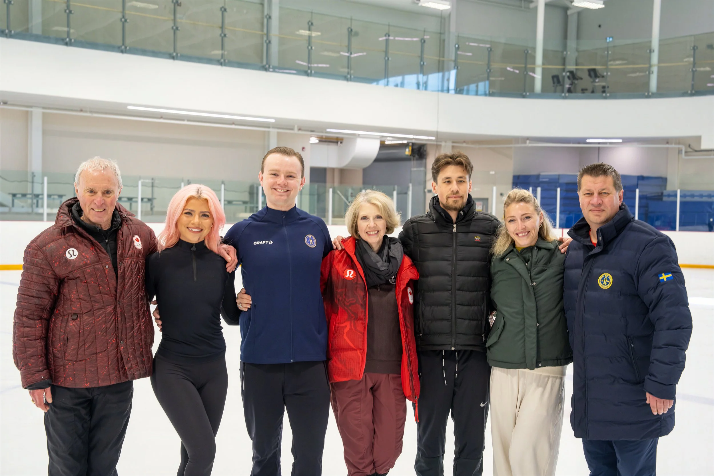 Group of seven people standing on an ice rink, posing for a photo, wearing winter athletic clothing, with a modern indoor ice rink facility in the background.