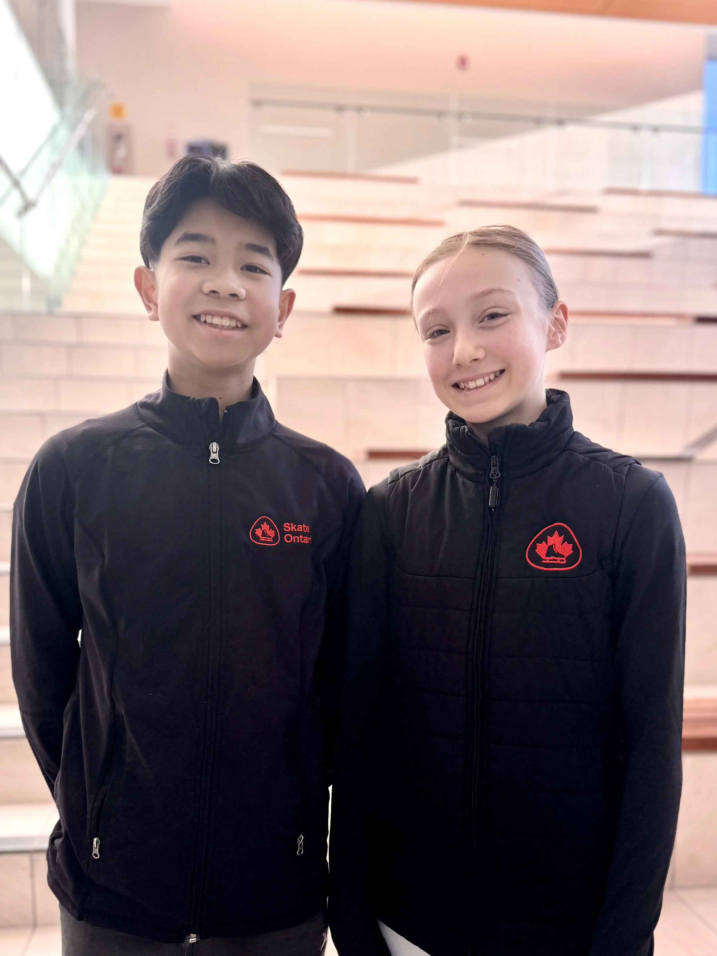 Two young children wearing black jackets with the Ontario Skate logo, smiling indoors against a background of stairs and a glass railing.