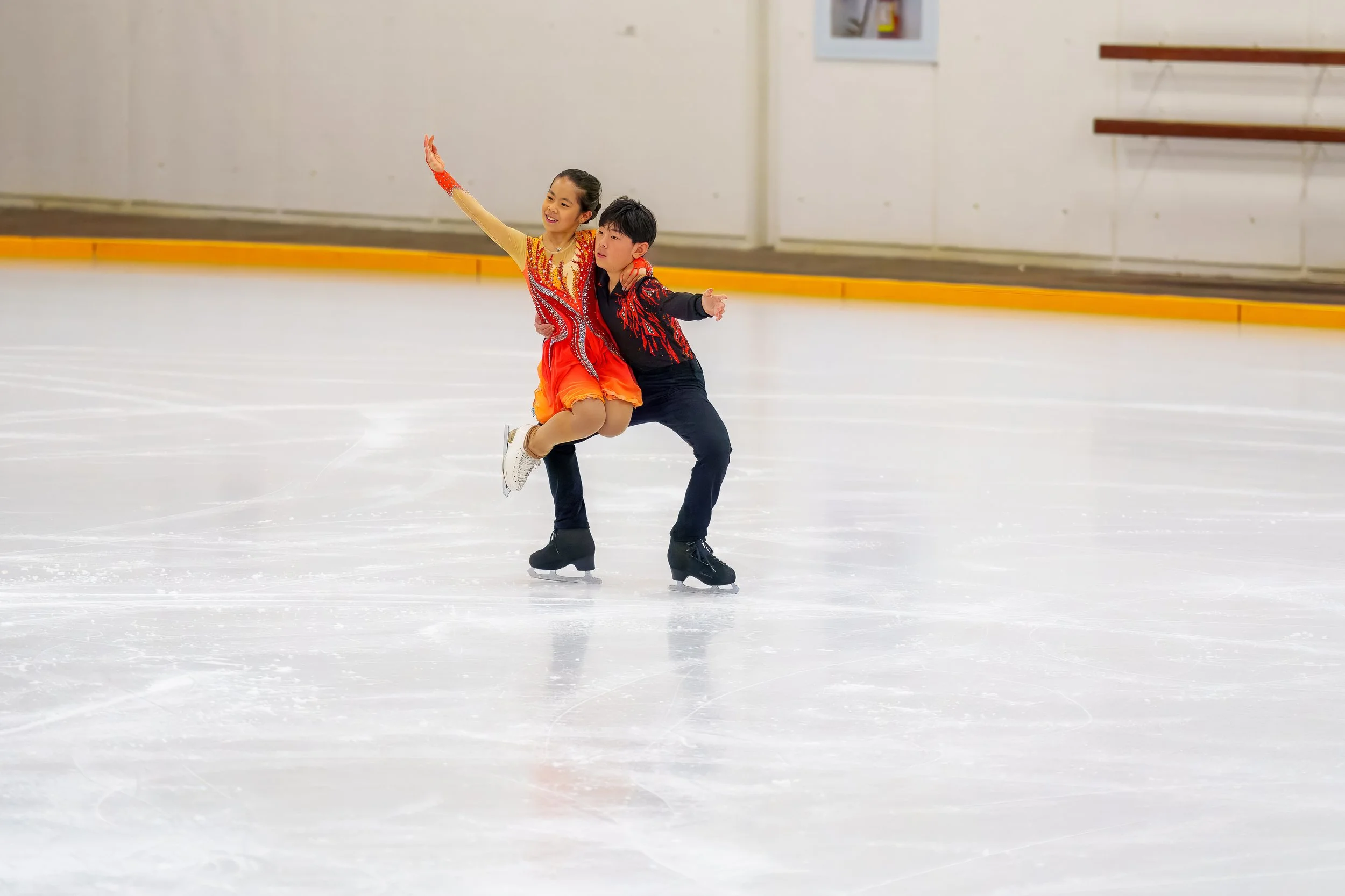 Two young figure skaters, a girl and a boy, performing together on an ice rink. The girl is mid-spin, smiling, with one arm raised and wearing an orange and red costume. The boy is supporting her, holding her around the waist, wearing a black and red costume. The background shows an indoor ice skating rink with a yellow barrier along the ice's edge.