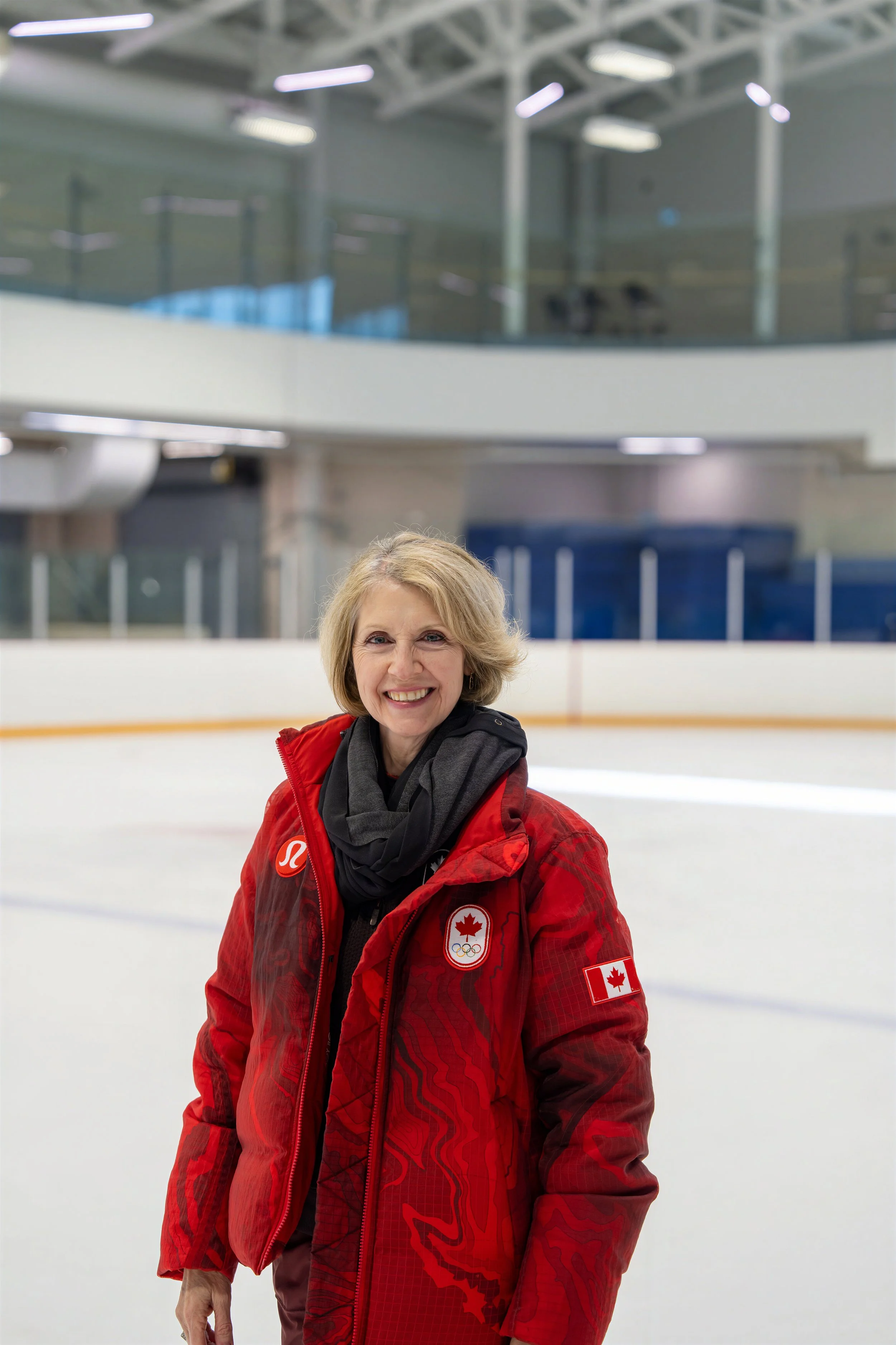 A woman in a red Canada Olympic jacket smiling on an indoor ice rink.