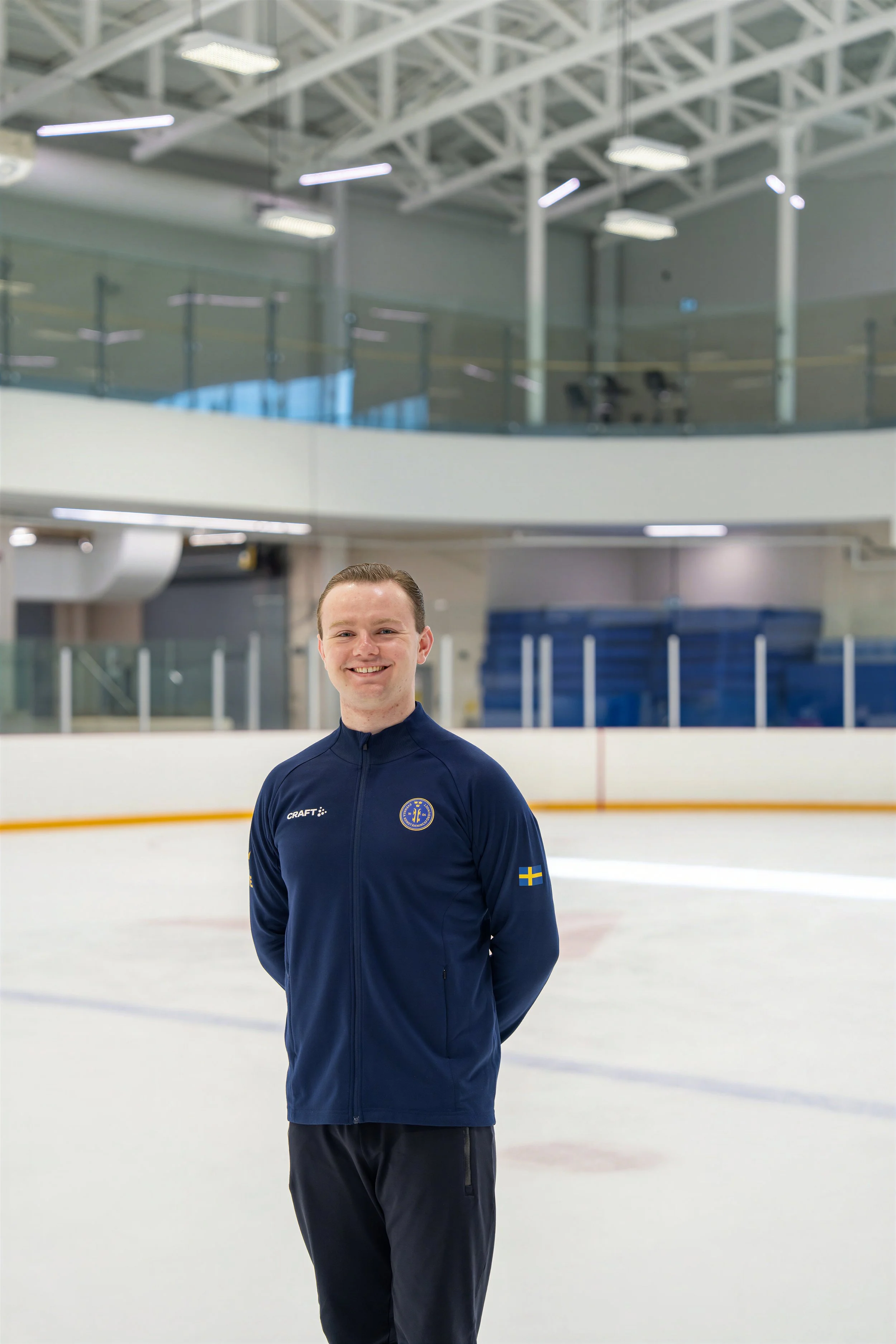 A young man in a navy blue sports jacket with Swedish flag patches on the shoulders, standing on an indoor ice rink with a blurred background of the rink and arena.