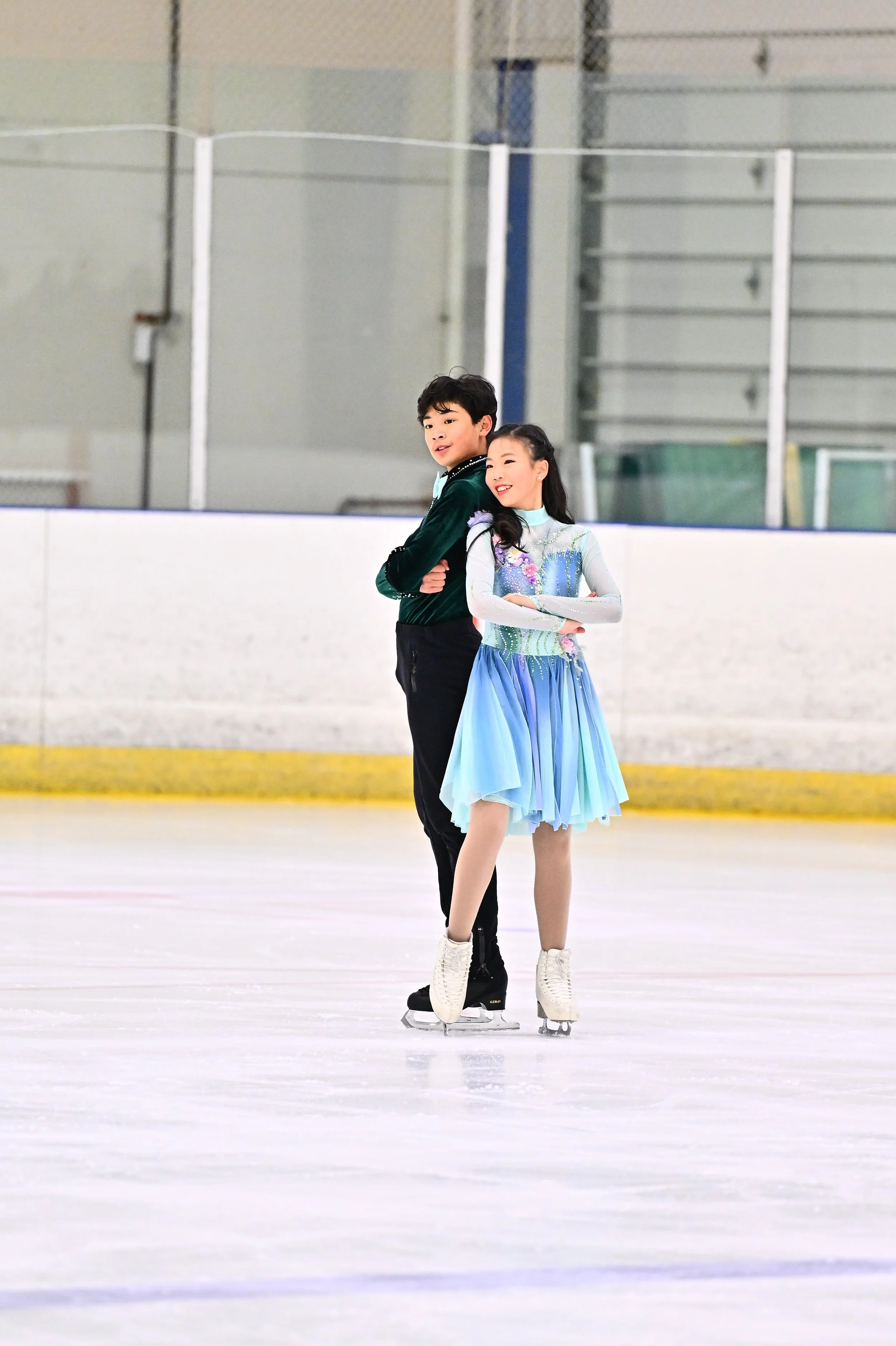 Two young figure skaters, a boy and a girl, standing on the ice rink. The girl is wearing a colorful dress with floral details, and the boy is dressed in a dark green jacket and black pants. They are both smiling and crossing their arms, posing for the photo.