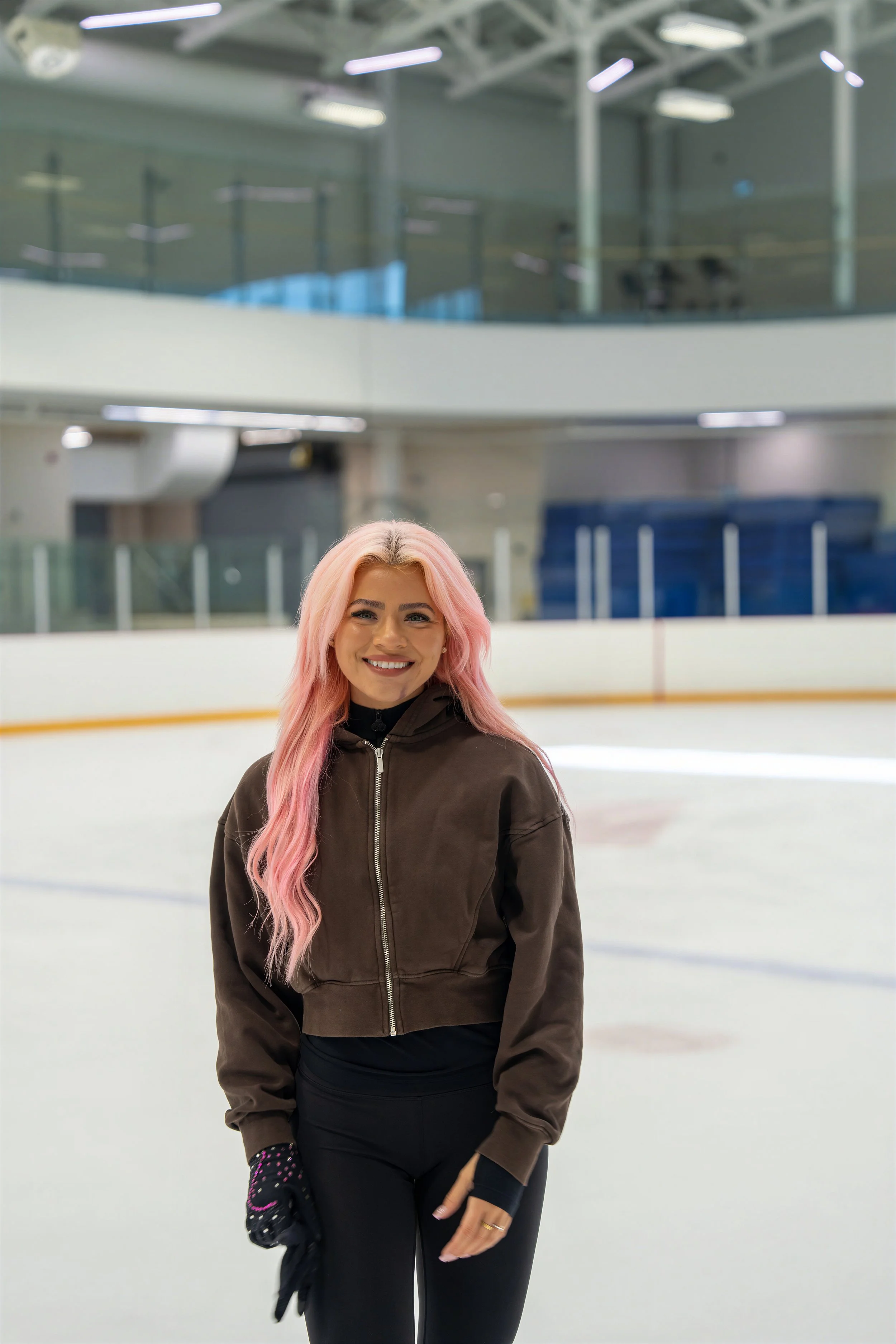 A woman with long pink hair smiling at an indoor ice skating rink.