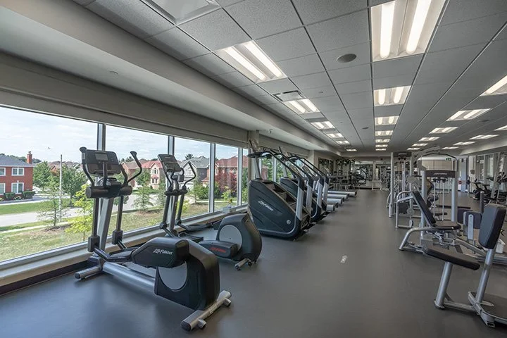 Empty gym with exercise machines and treadmills along large windows with a view of houses and trees outside.