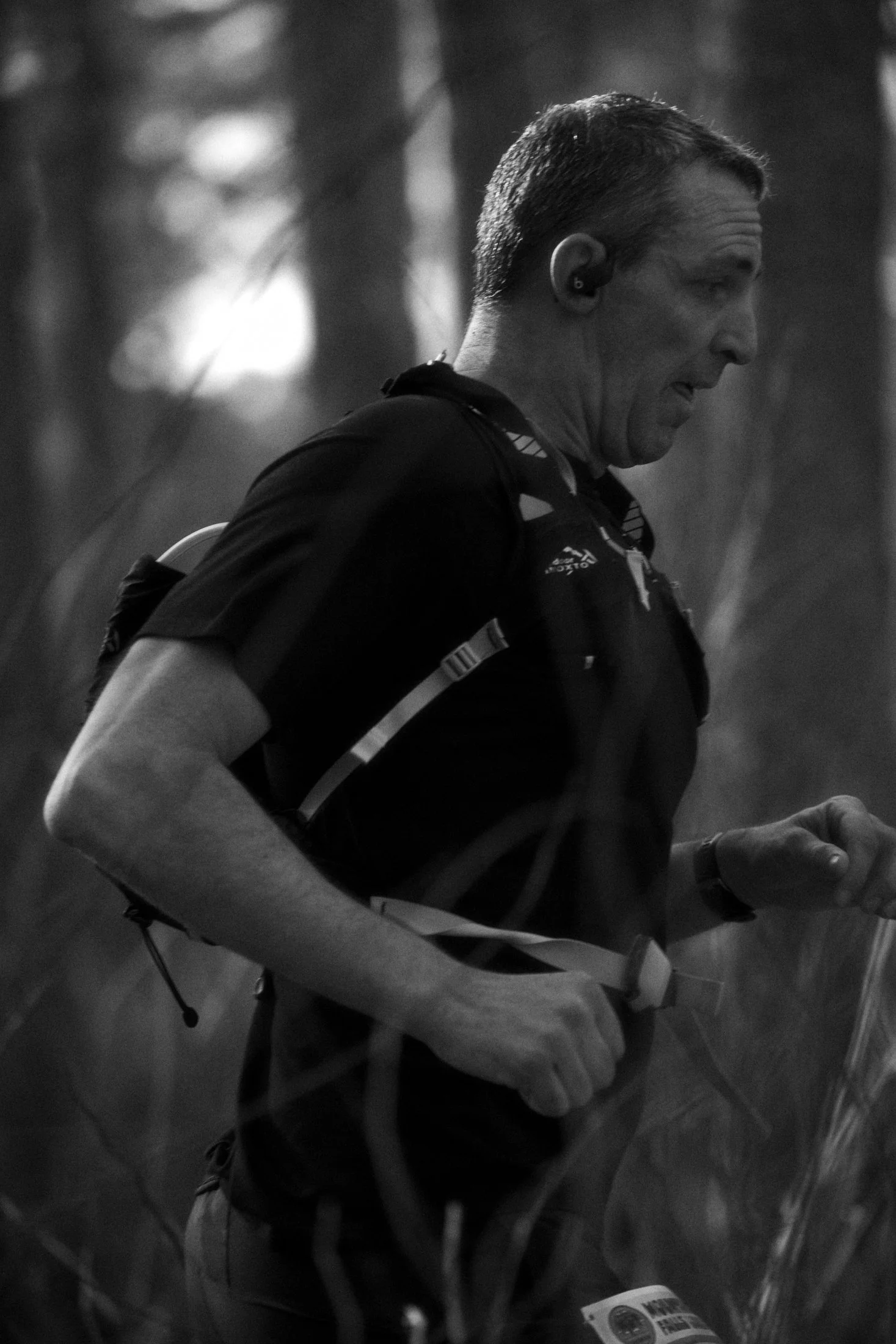 A man running through a wooded area wearing a black shirt, a backpack, and earbuds.