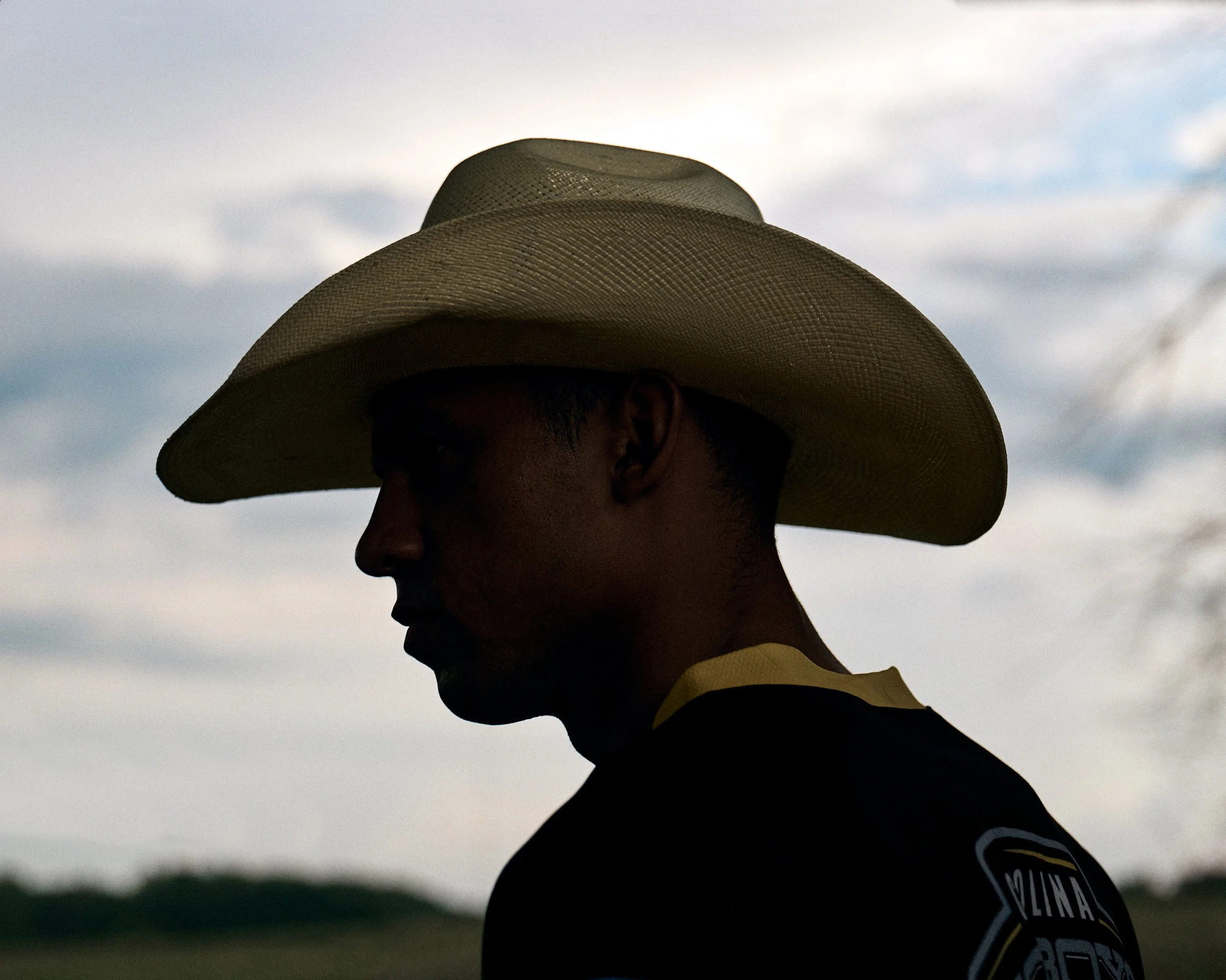 Silhouette of a person wearing a wide-brimmed hat, facing left with a cloudy sky in the background.