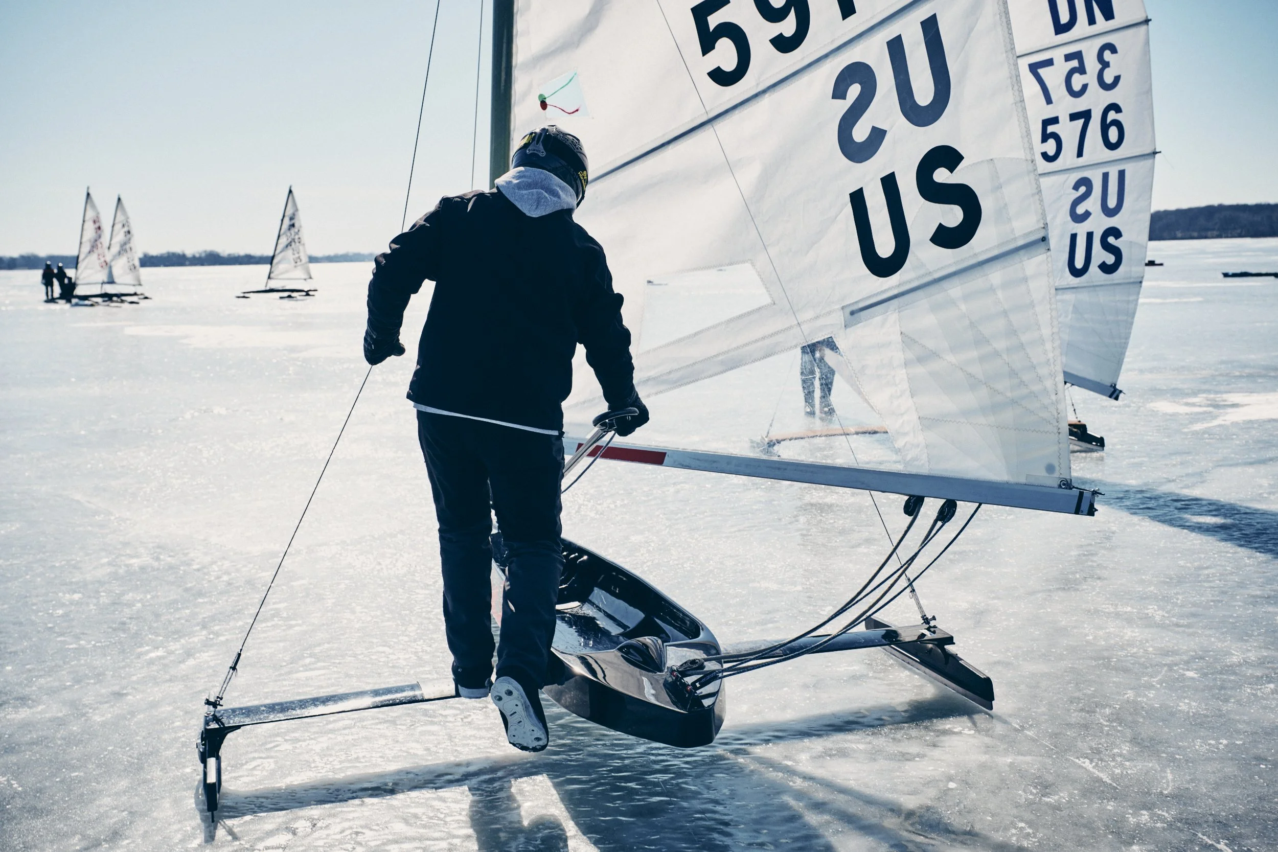 Person standing on ice in a snowsled, holding onto the side, with ice sailboats in the background on a frozen lake.
