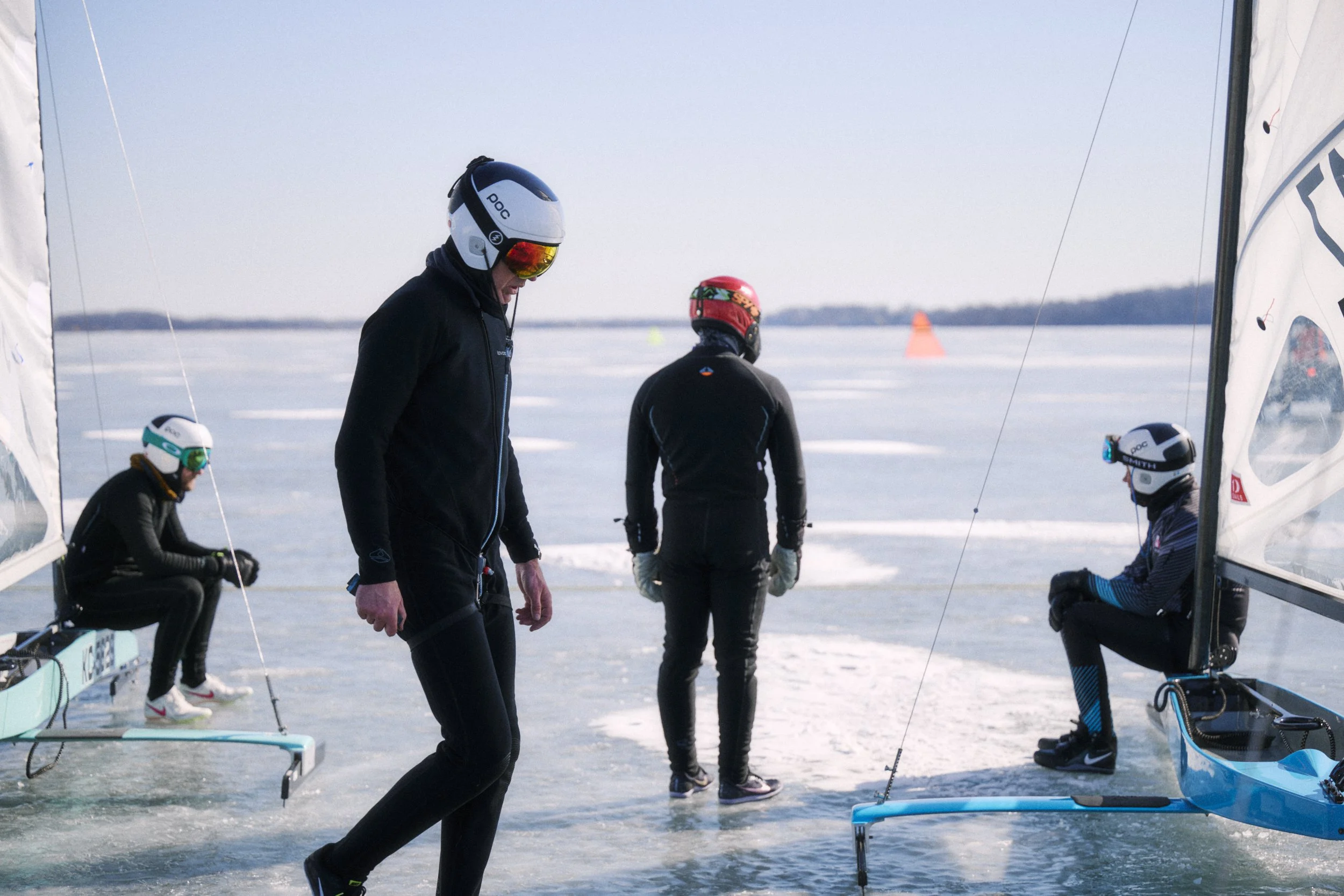 Four people preparing ice boats on a frozen lake, wearing helmets and winter gear, with orange markers in the background.