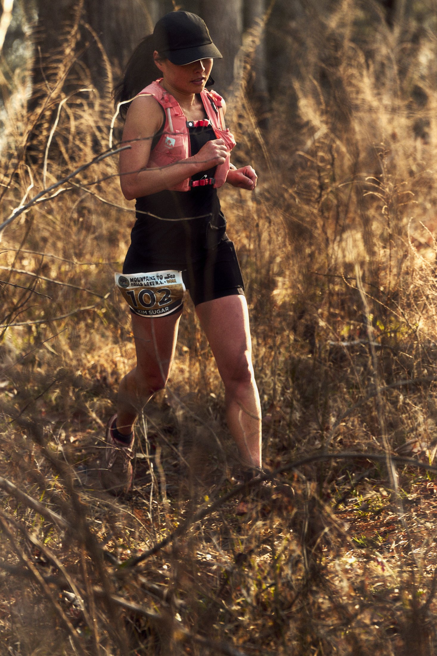 A female trail runner wearing black shorts, a black cap, and a pink vest with a bib number 102 runs along a dirt path through dry, brown vegetation in a natural outdoor setting.