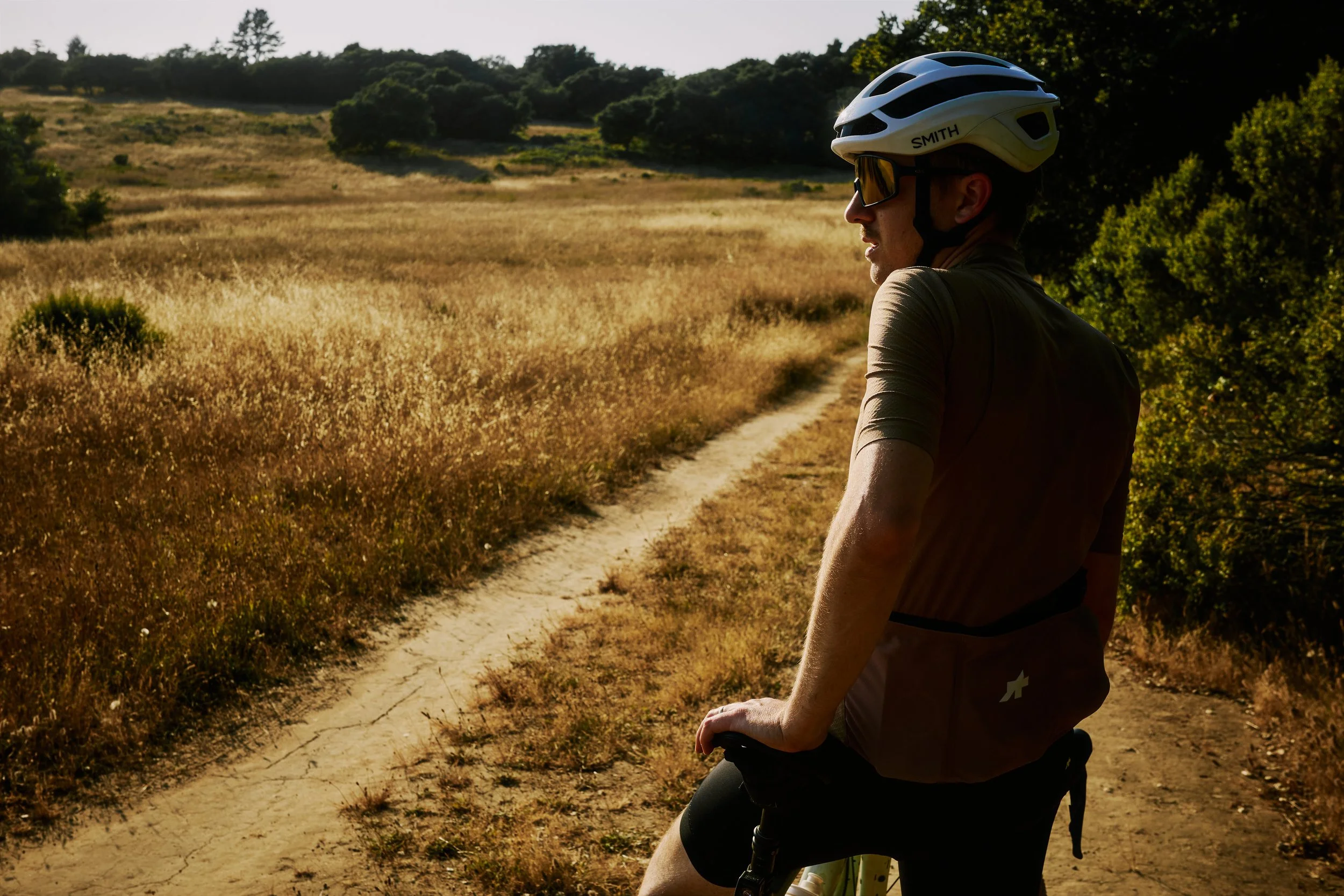 A person wearing a white bike helmet, sunglasses, and a brown cycling outfit riding a bike along a dirt trail in a countryside with dry grass and trees.