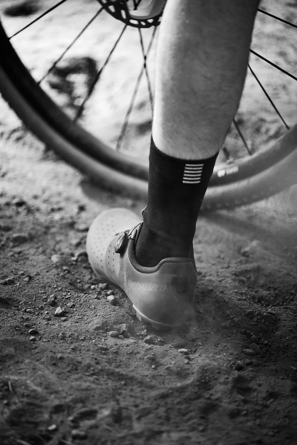 Close-up of a person's foot in a cycling shoe, wearing a sock, next to a bicycle tire on a dirt trail.