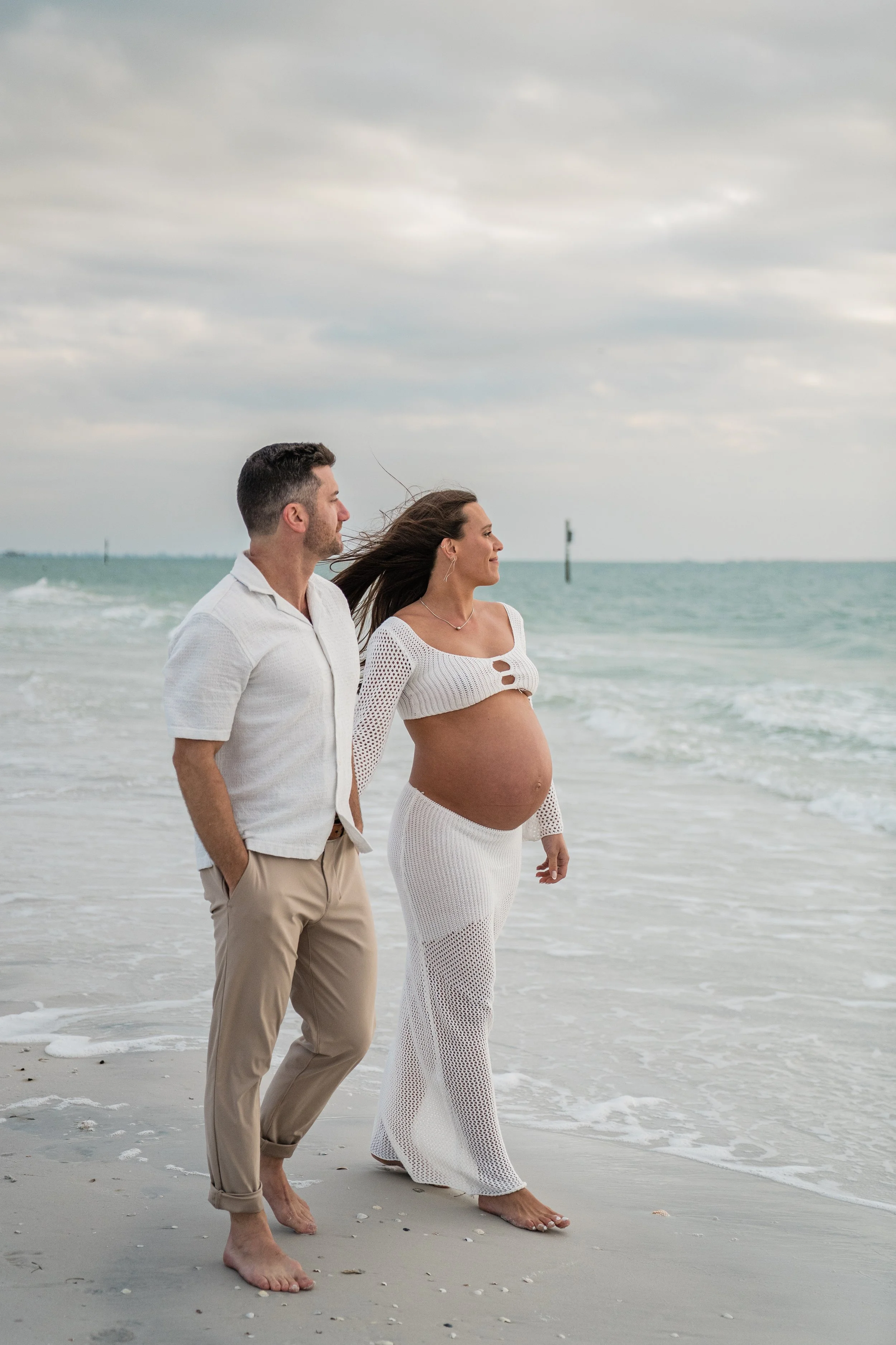 A pregnant woman and a man walking barefoot on a beach, looking at the ocean under cloudy skies.