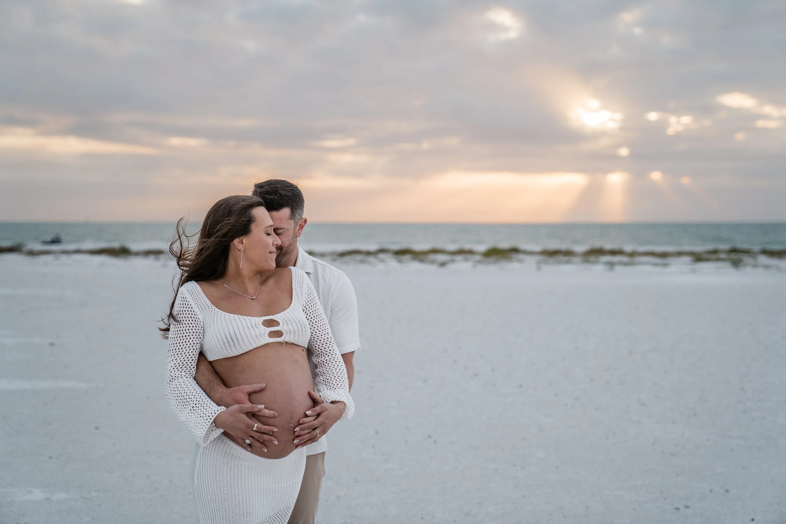 A pregnant woman and her partner embrace on a beach during sunset, with the woman wearing a white crochet top and skirt, and the man standing behind her with arms around her belly.