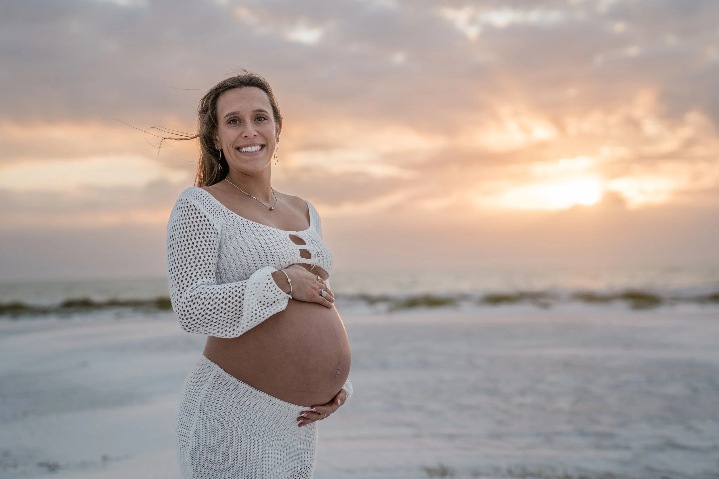 A pregnant woman in a white crochet top and skirt standing on the beach during sunset, smiling and cradling her belly.