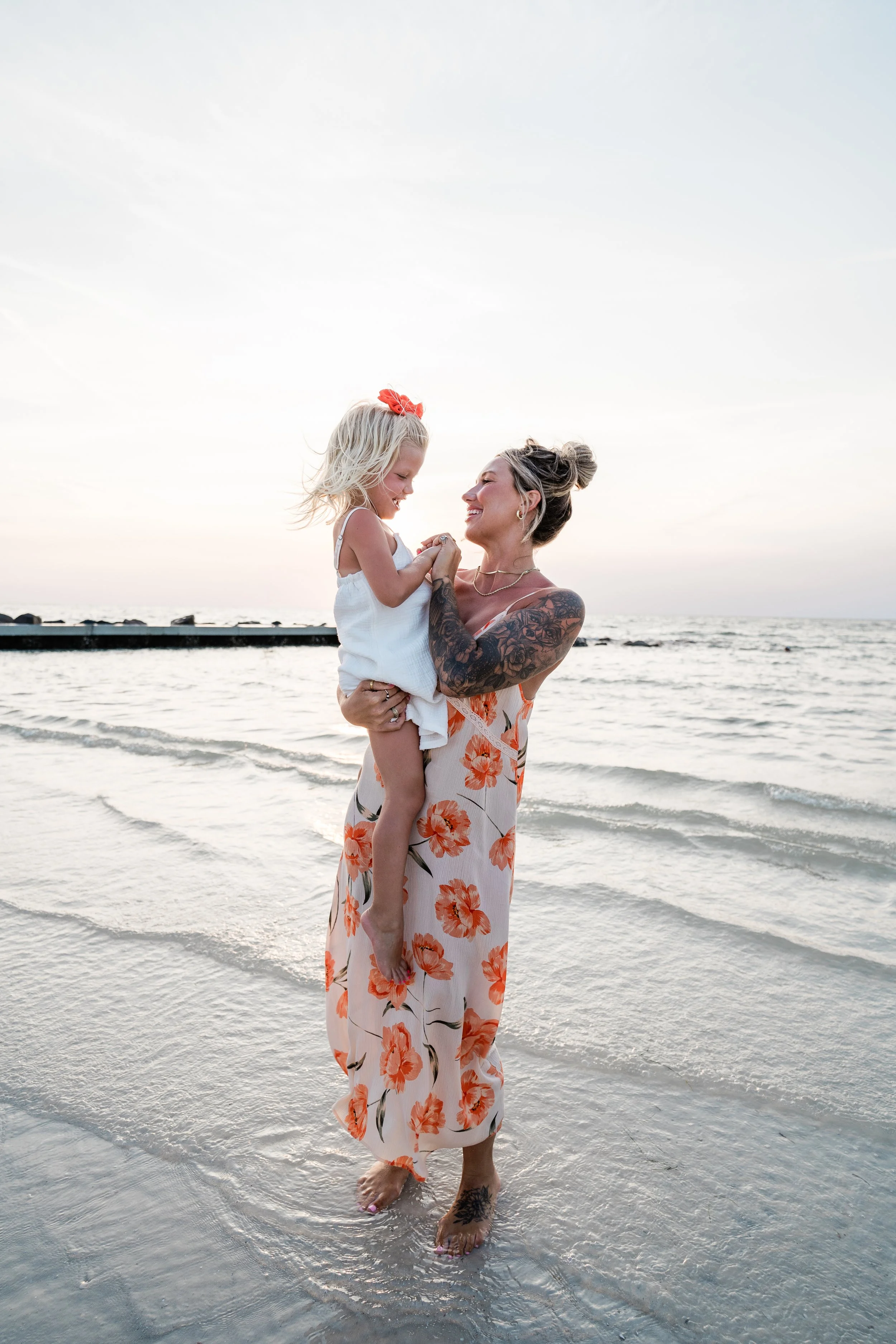 A woman with tattoos wearing a floral dress holds a young girl in a white dress at the beach during sunset, smiling and enjoying their time.