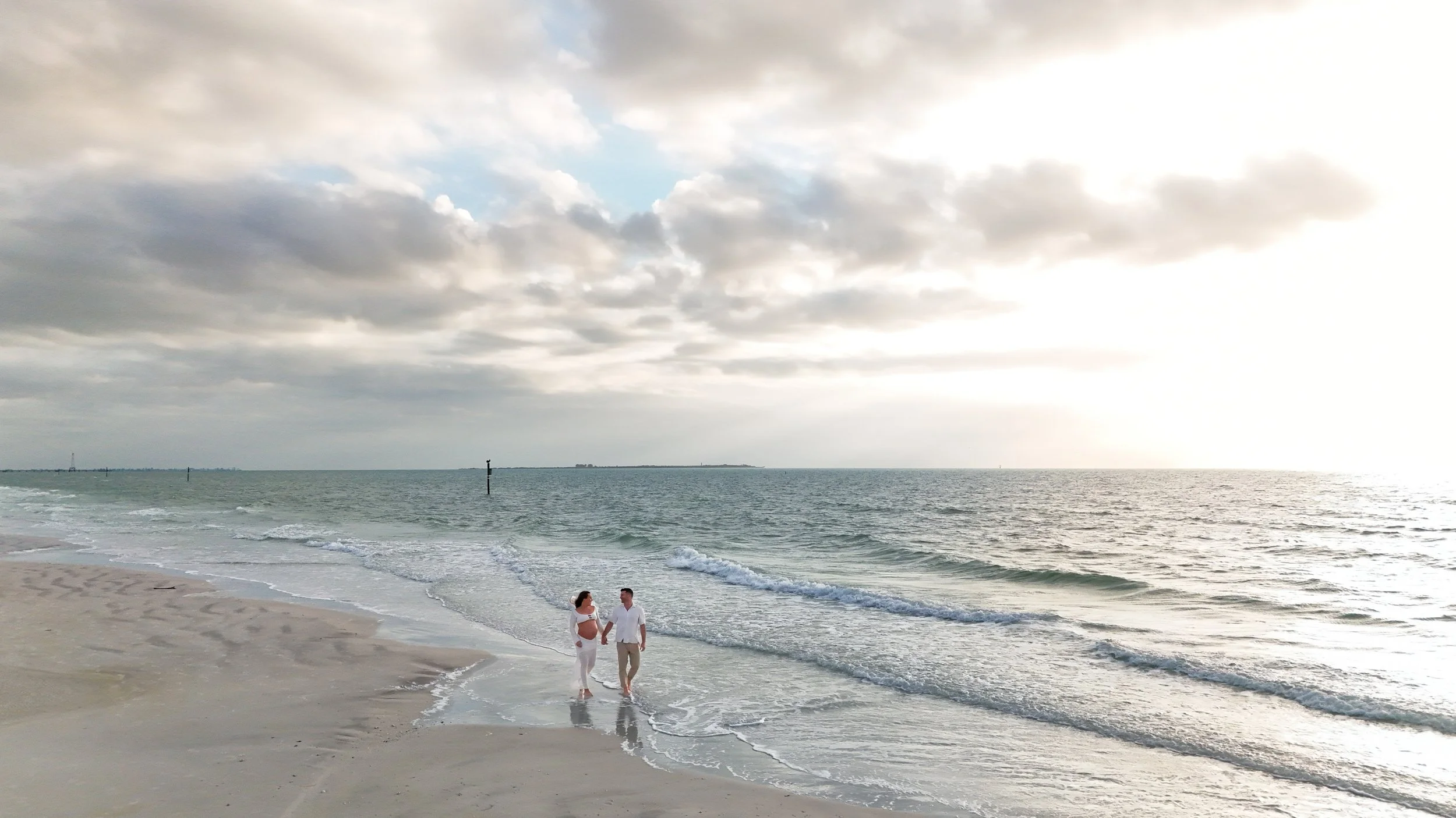 A couple holding hands walking along the shoreline of a beach with cloudy sky and calm ocean waves