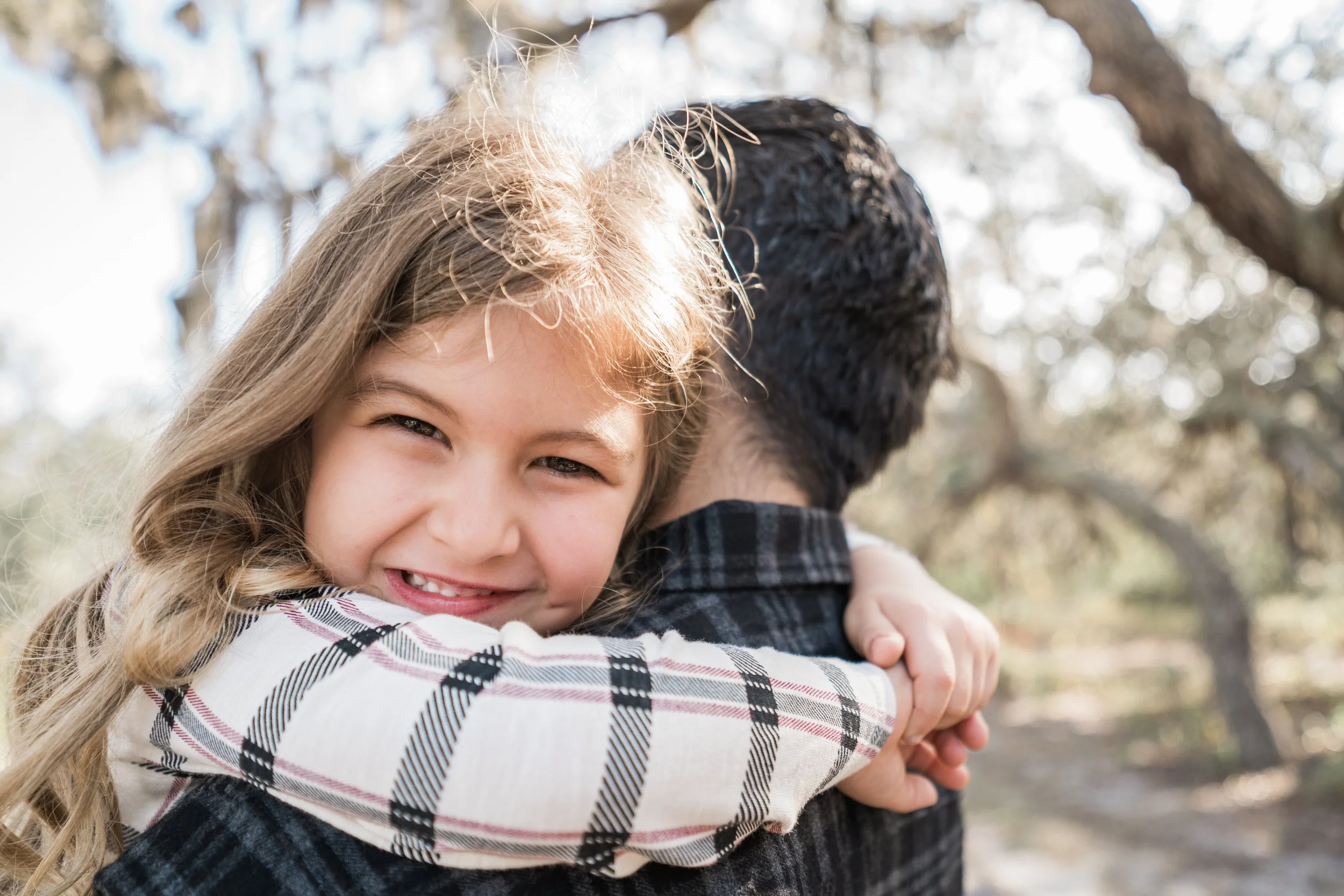 A young girl with long blonde hair hugging a man from behind, smiling at the camera, outdoors with trees in the background.