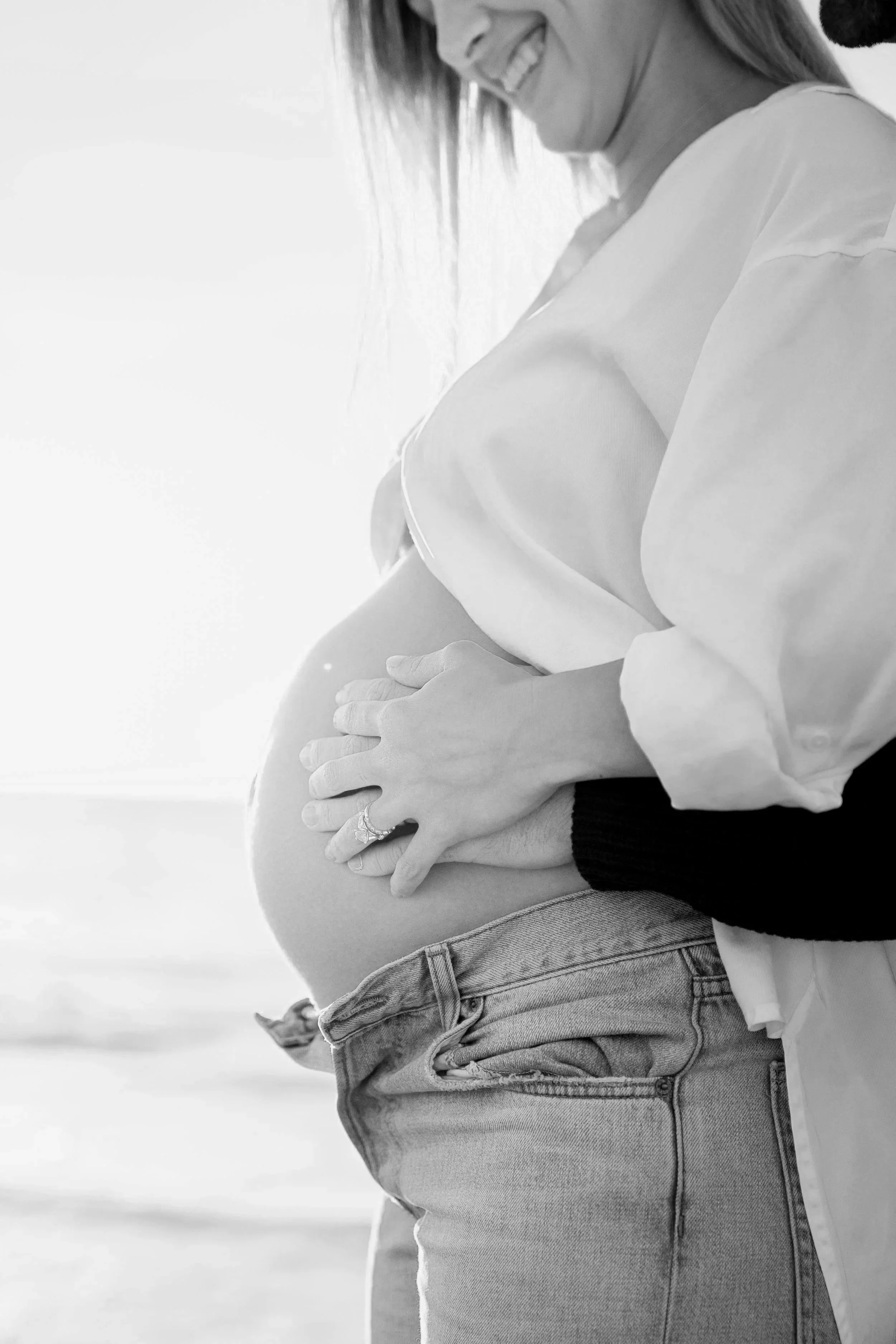Black and white photo of a pregnant woman smiling and holding her belly, wearing a loose shirt and jeans.