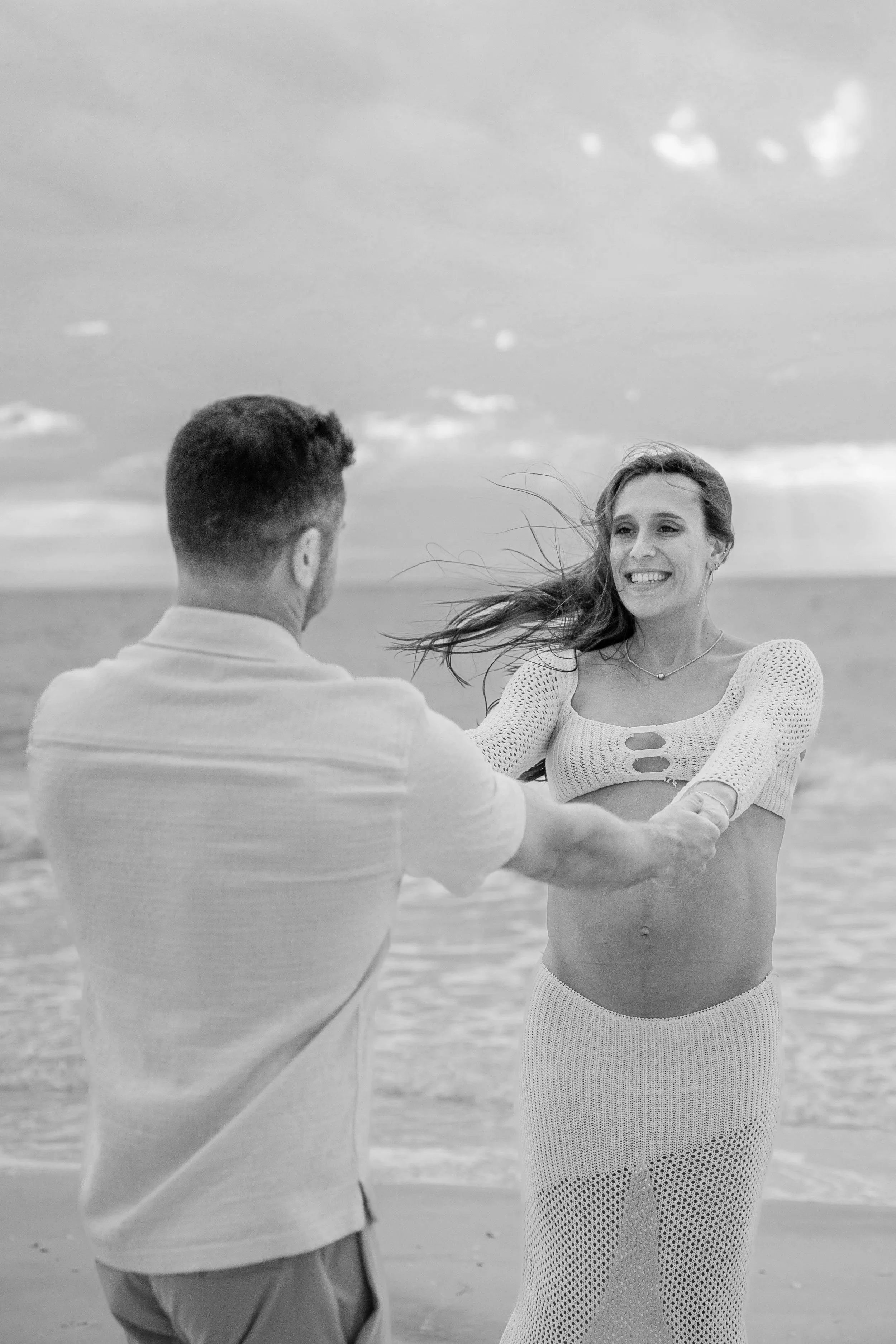 A black-and-white photo of a couple holding hands on the beach, with the woman smiling and the man facing away from the camera, the woman has long hair and is wearing a knitted top and a skirt, and the sky and ocean are visible in the background.