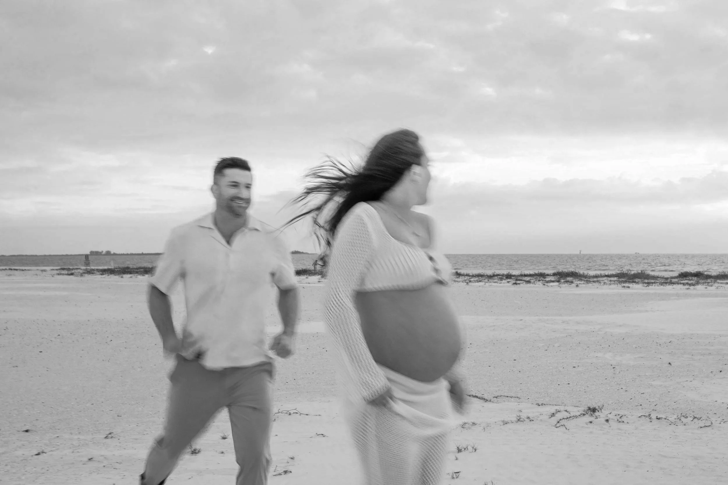 A woman with a pregnant belly walking on the beach while a man runs towards her, both smiling, in black and white.