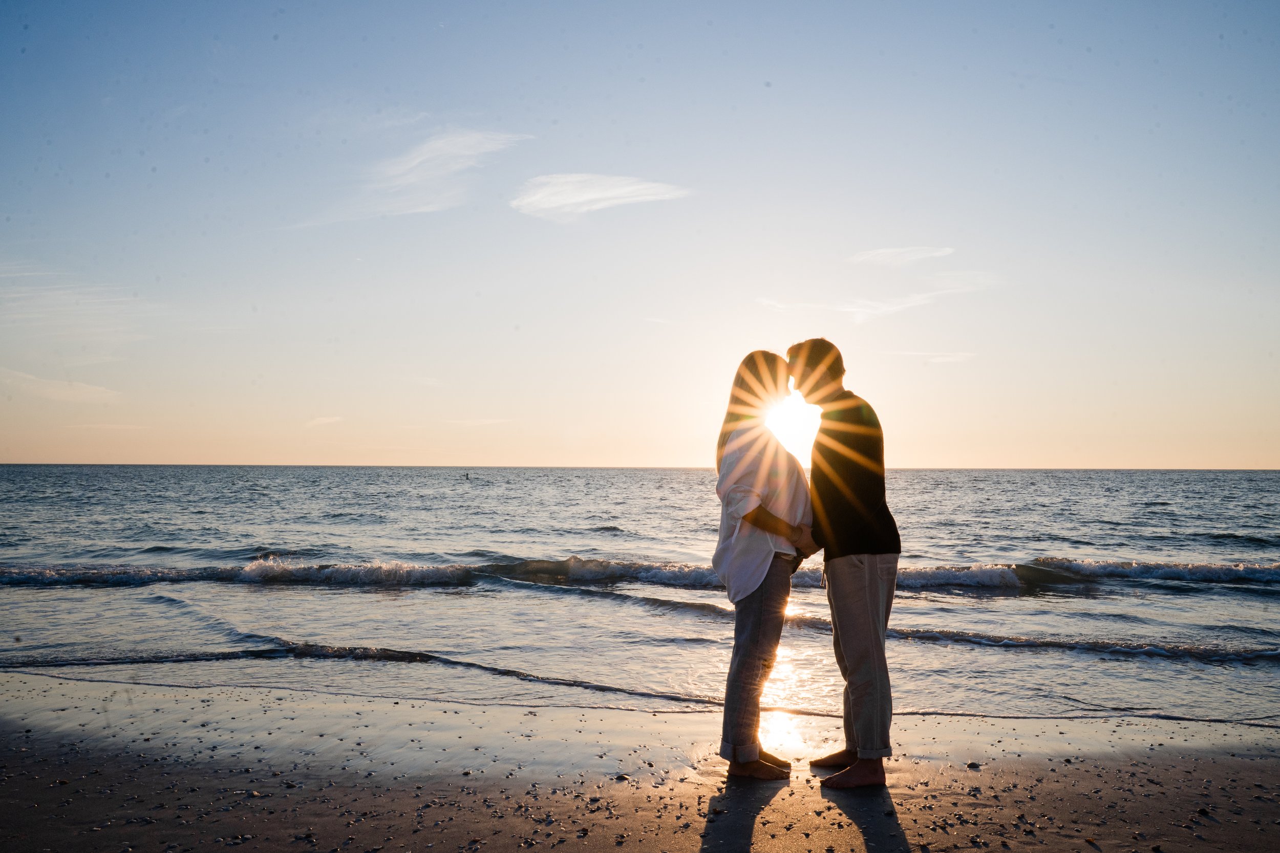A couple on the beach at sunset, kissing with their foreheads touching, the sun shining through their faces creating a starburst effect.