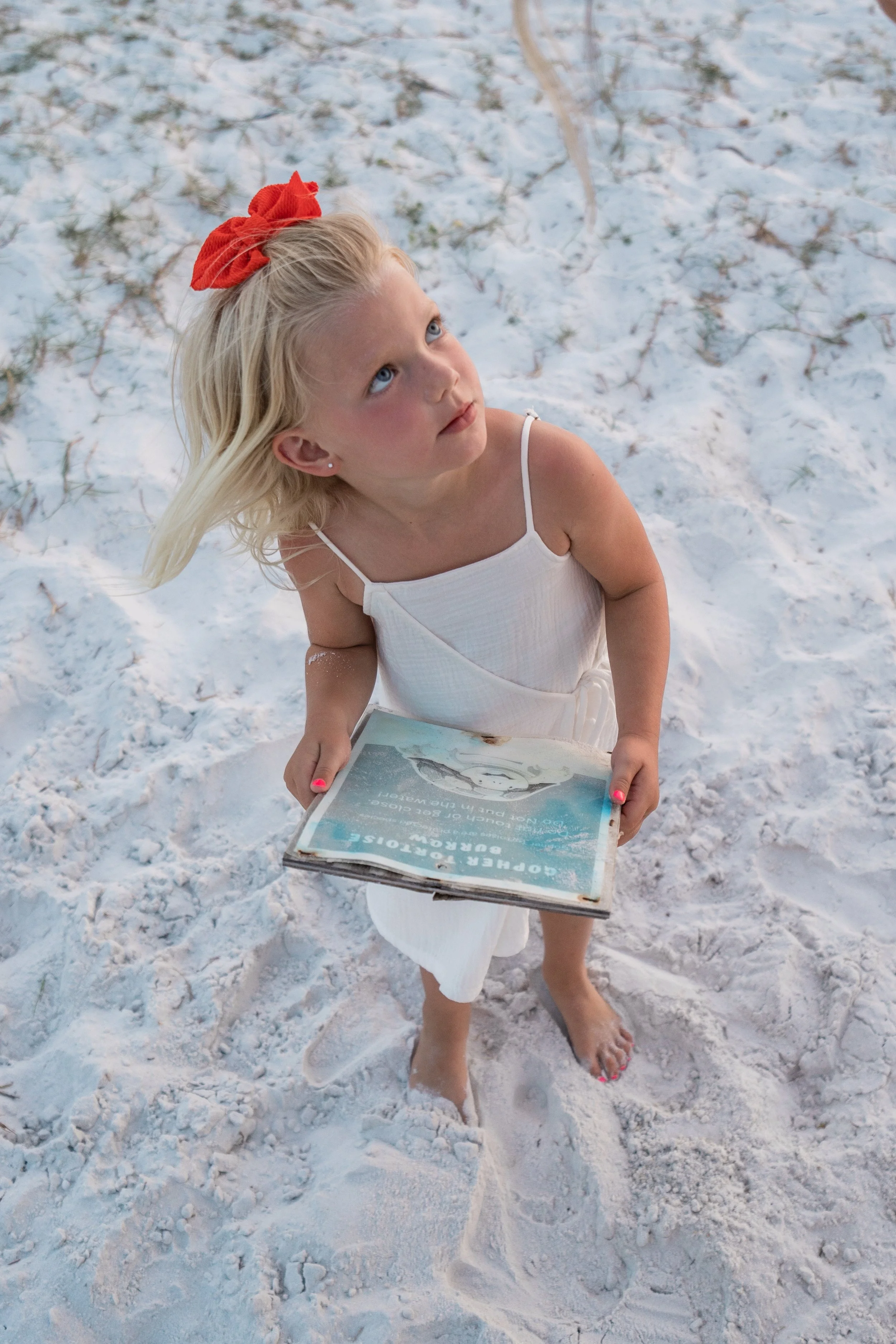 A young girl with blonde hair, a red bow, and a white dress, standing barefoot in the sand, looking up, holding a magazine titled 'Goper Bouteose' with an image of an iceberg on the cover.