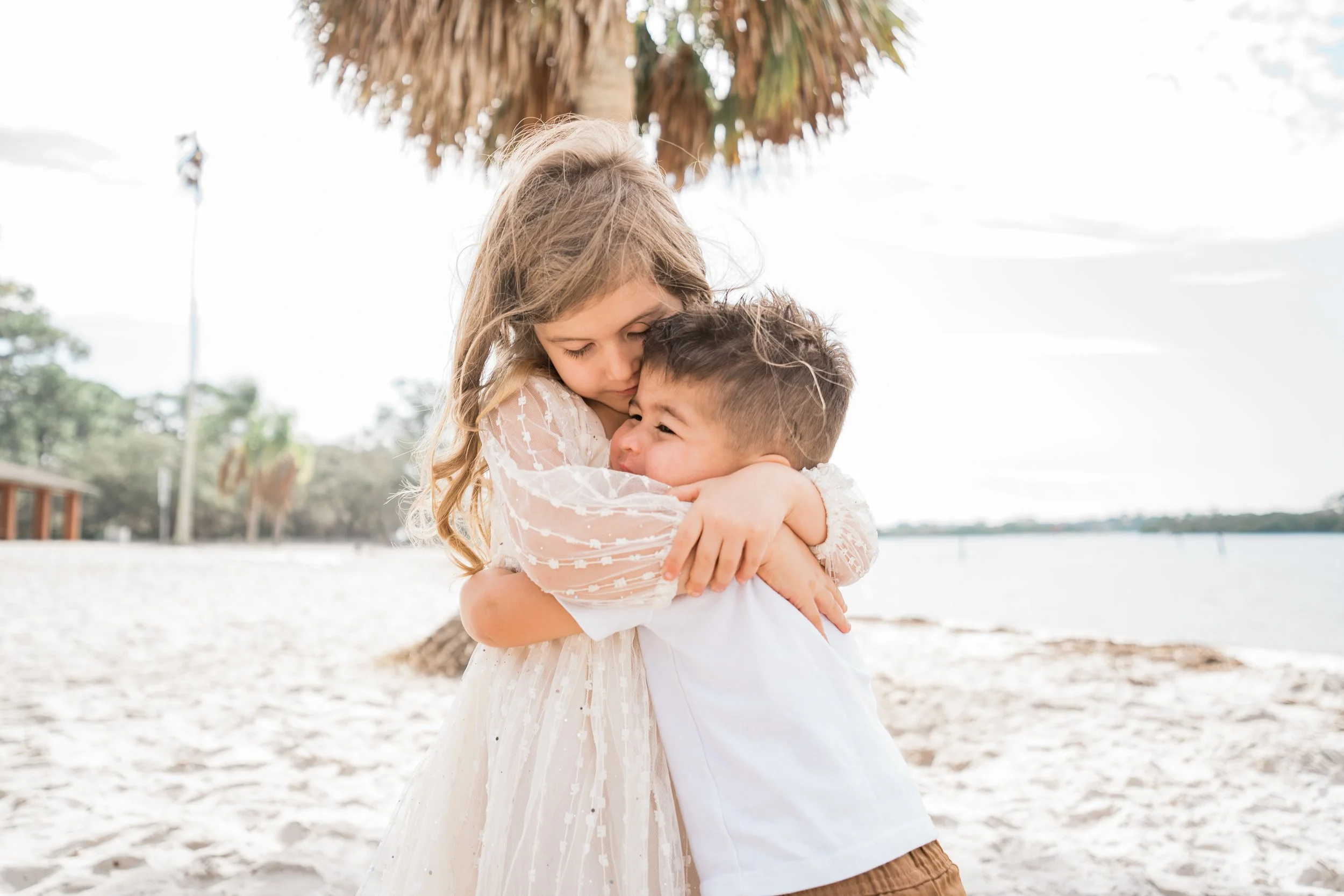 A young girl hugging a boy in front of a beach scene with a palm tree, water, and cloudy sky.