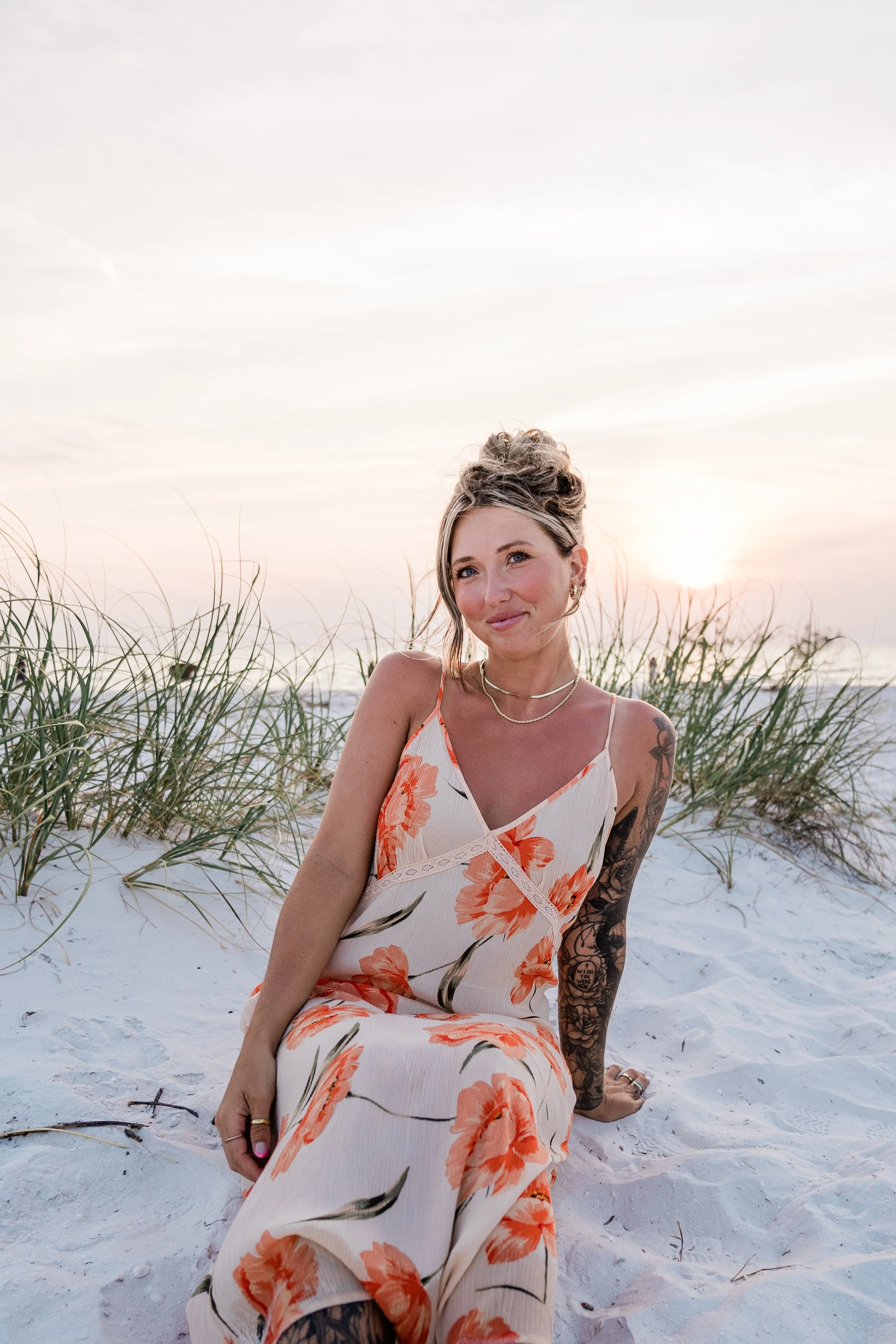 A woman with tattoos wearing a floral dress sitting on the white sand at the beach during sunset.