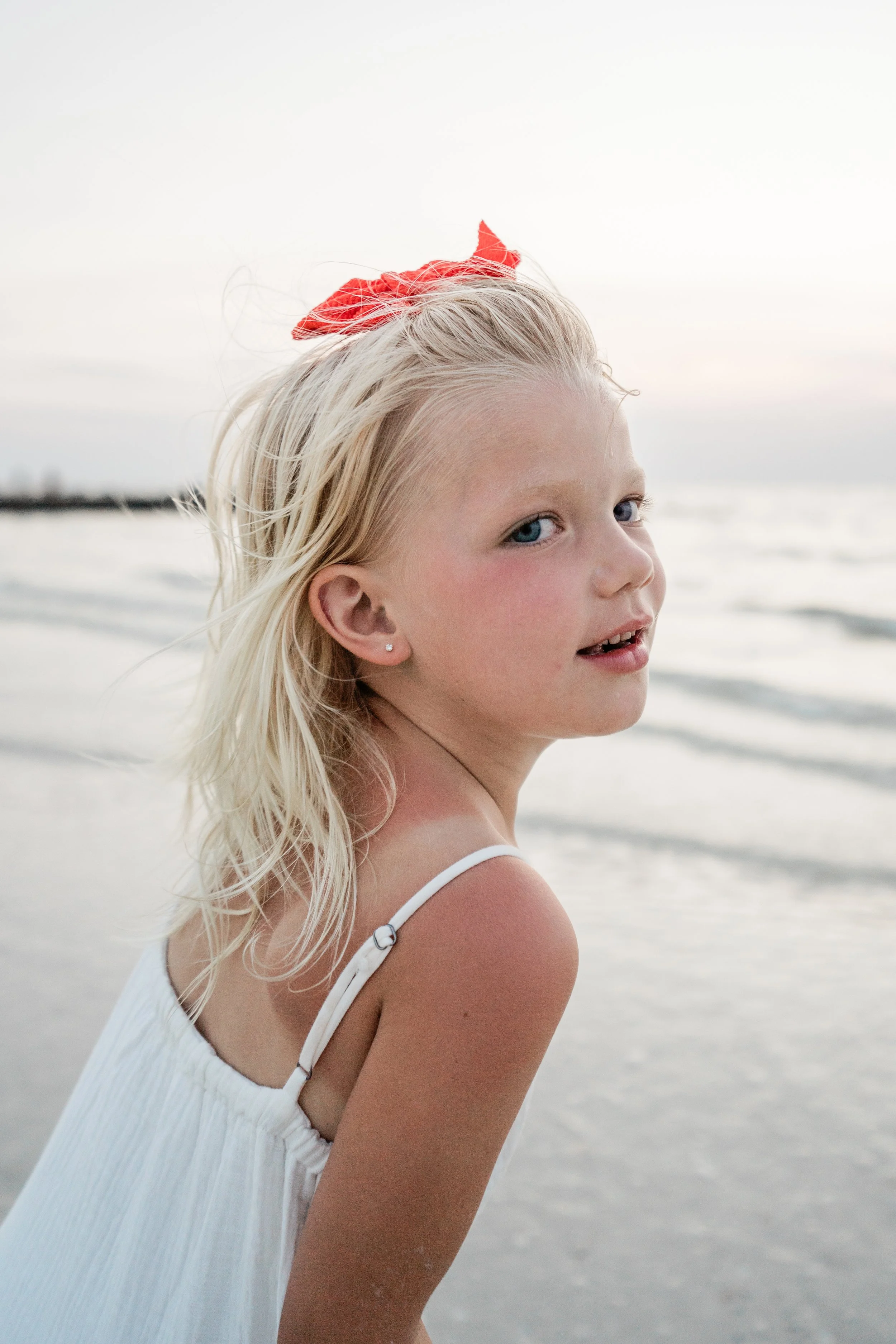 A young girl with blonde hair and blue eyes wearing a white dress and a red bow on her head, standing on the beach with the ocean in the background.