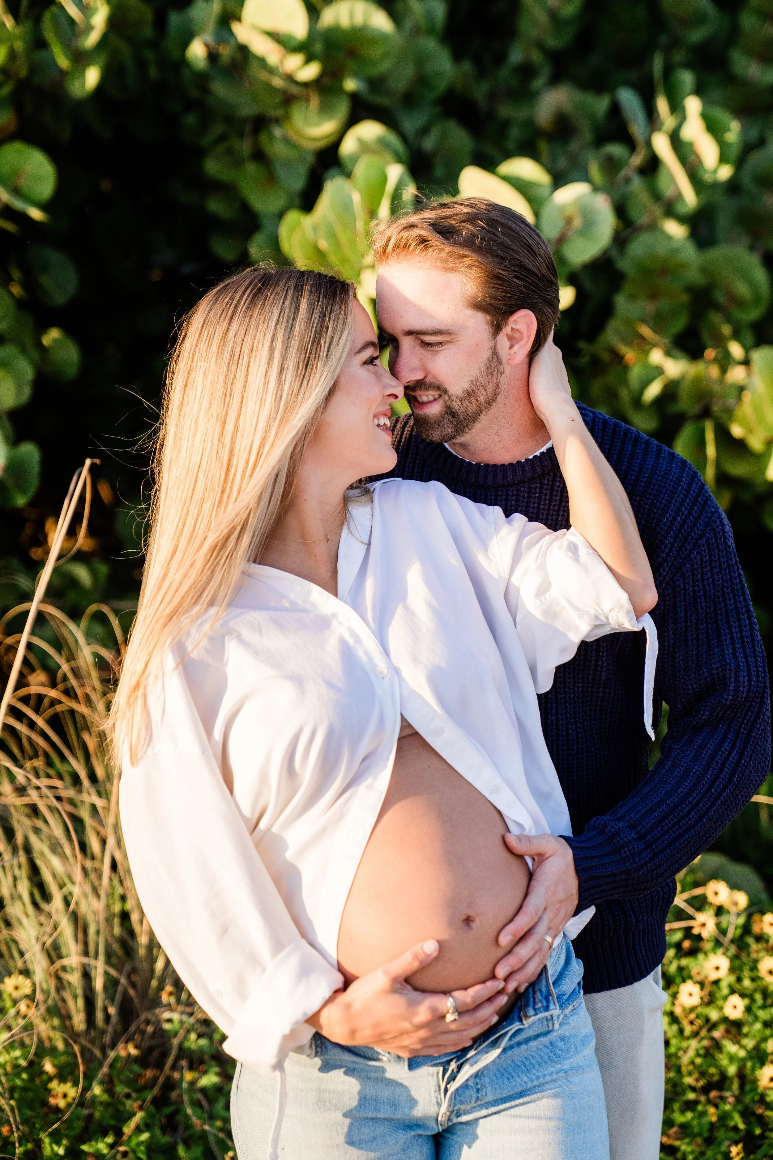 A pregnant woman and a man are outdoors, close to each other, smiling and touching her belly. The woman has blonde hair and is wearing a loose white shirt and jeans. The man has brown hair, a beard, and is wearing a dark blue sweater. They are surrou