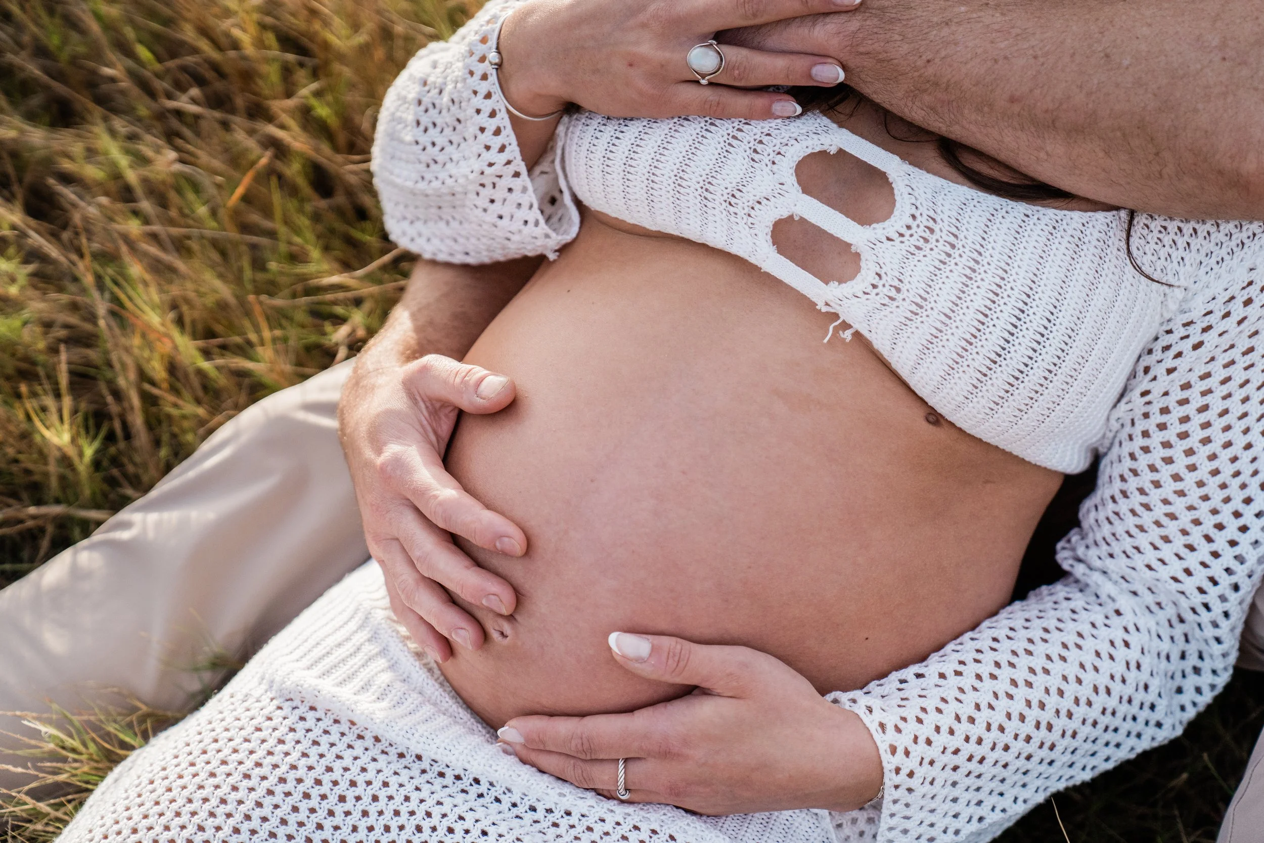 Close-up of a pregnant woman's belly with her partner's hands gently holding it, outdoors on grass, woman wearing a white crochet top.