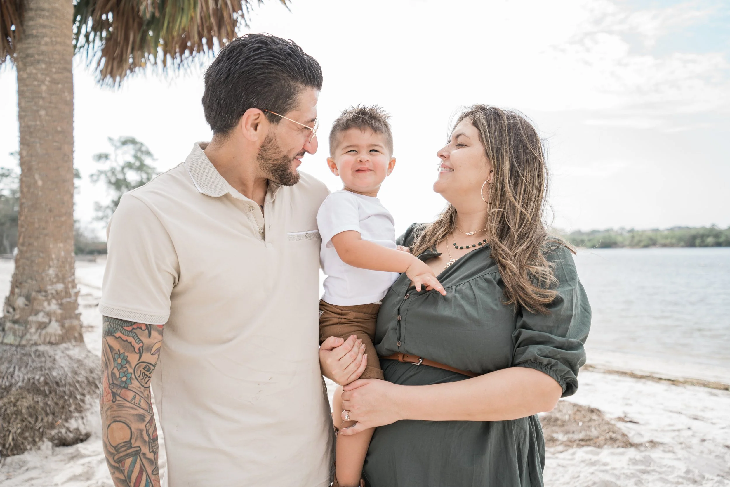 A happy family of three enjoying a day at the beach, with a man, woman, and young boy, standing near the water under a palm tree.