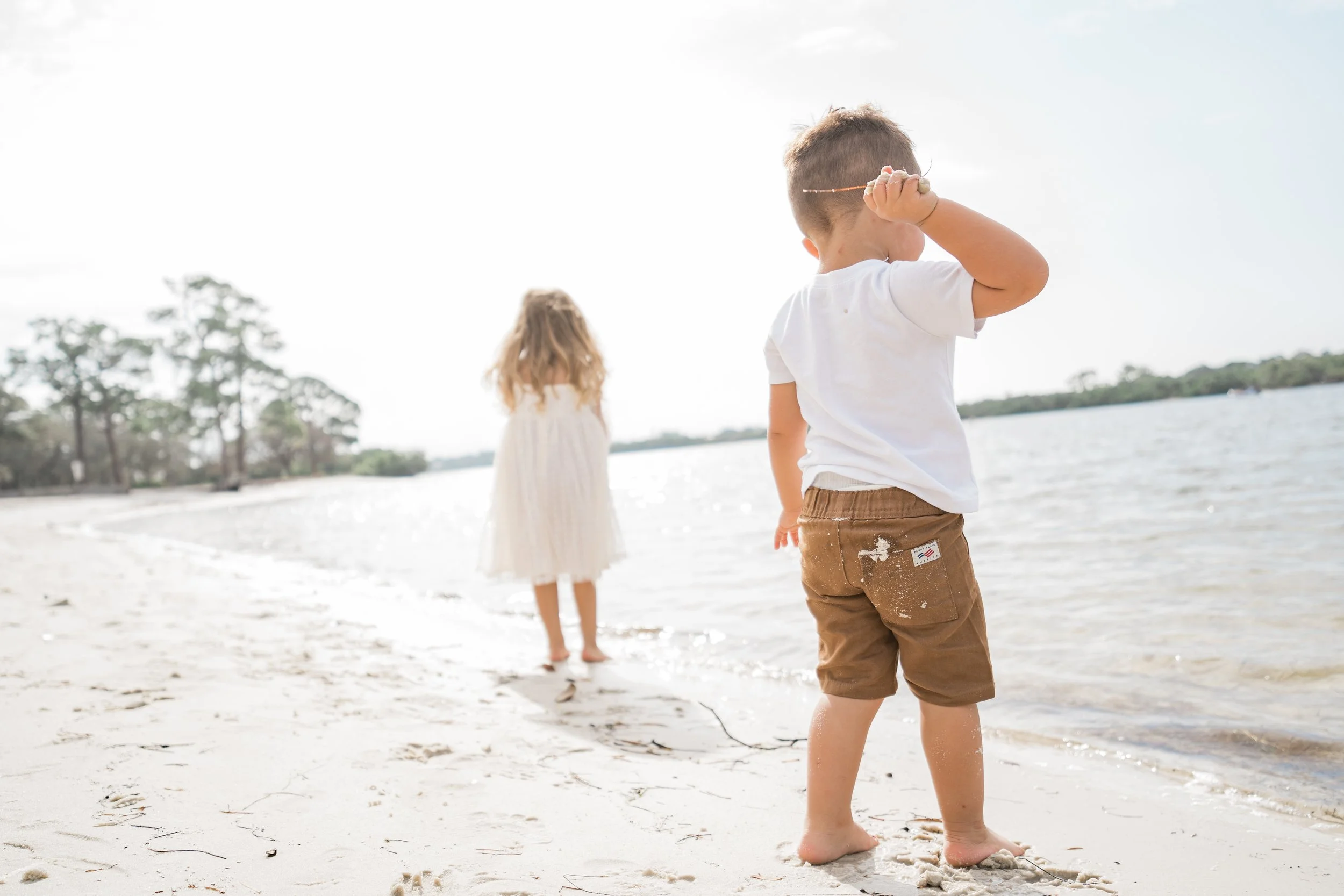 A young boy and girl playing on a beach near the water, with trees in the background and clear skies.
