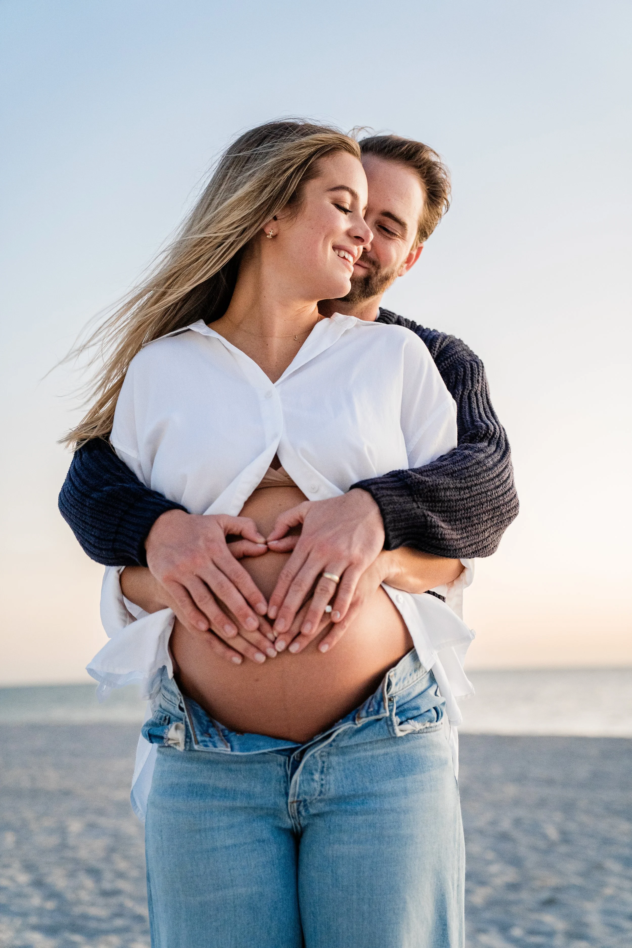 A couple stands on the beach, smiling, with the man embracing the woman from behind and both making a heart shape with their hands over her pregnant belly during sunset.