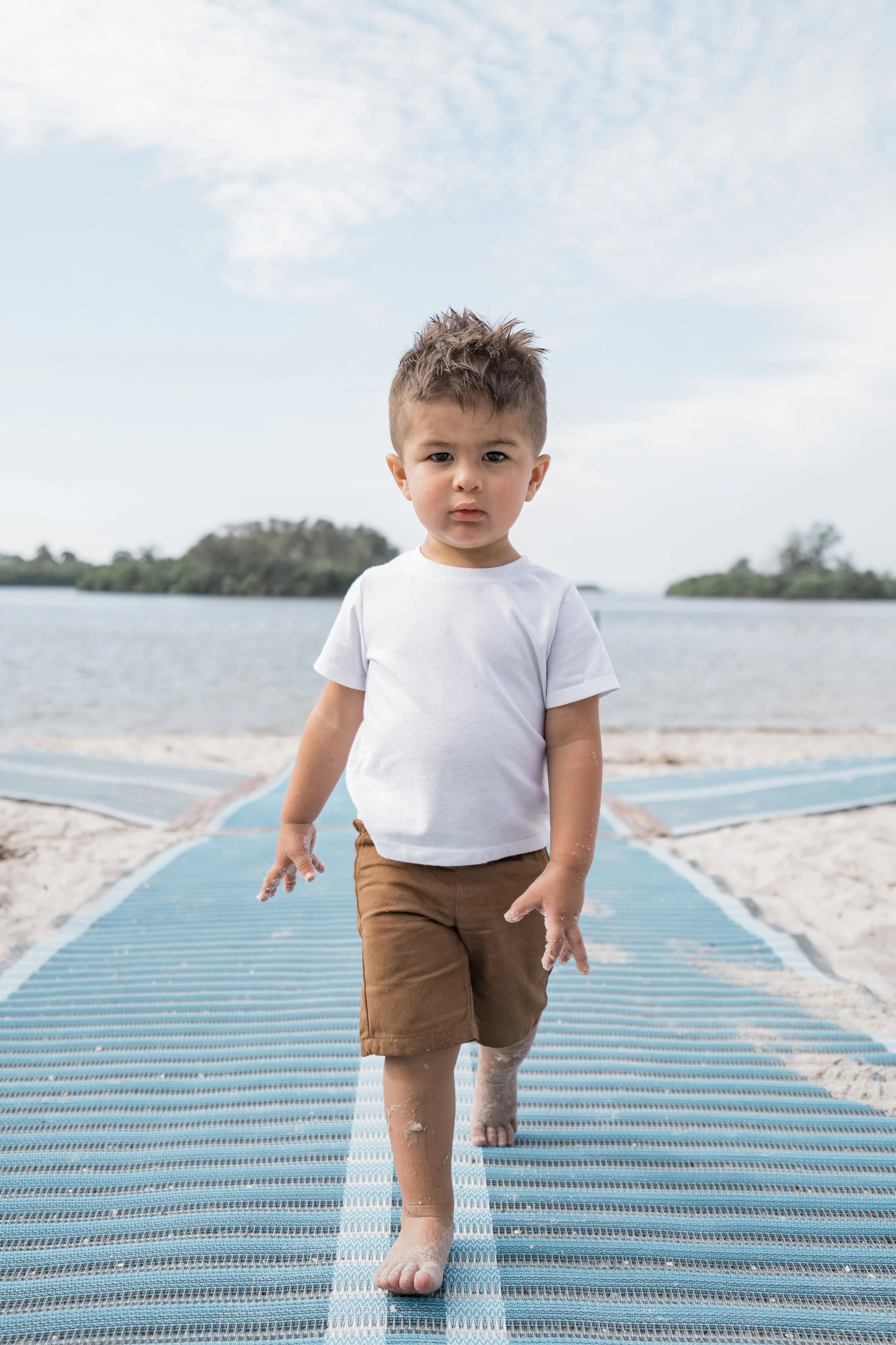 Young boy walking on a path at the beach, sand and water visible, with a cloudy sky overhead.